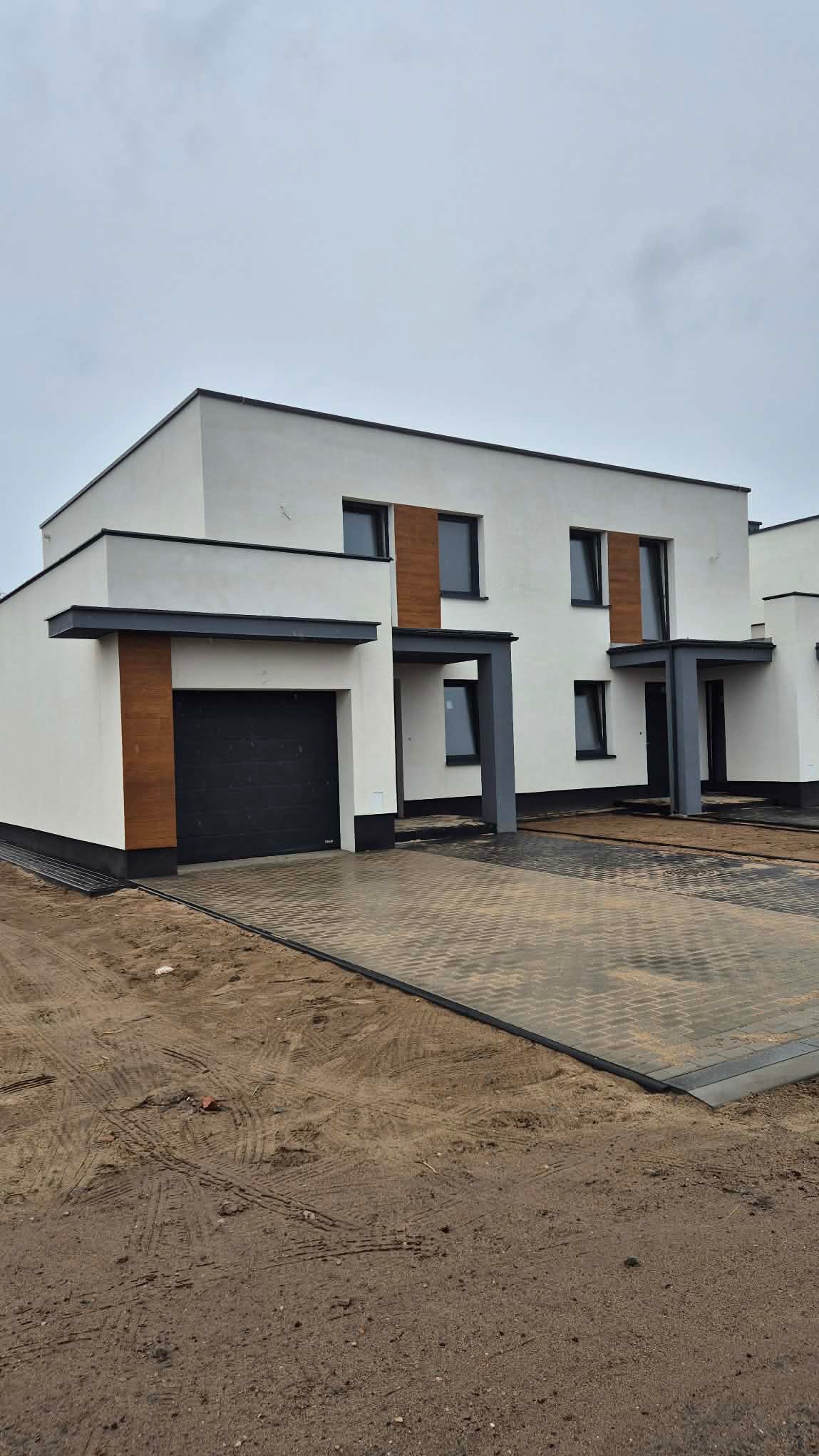 Nowoczesny dom bliźniak z garażem i elewacją w bieli, z elementami dekoracyjnymi w odcieniach drewna. Kostka brukowa przed budynkiem, pochmurne niebo w tle.