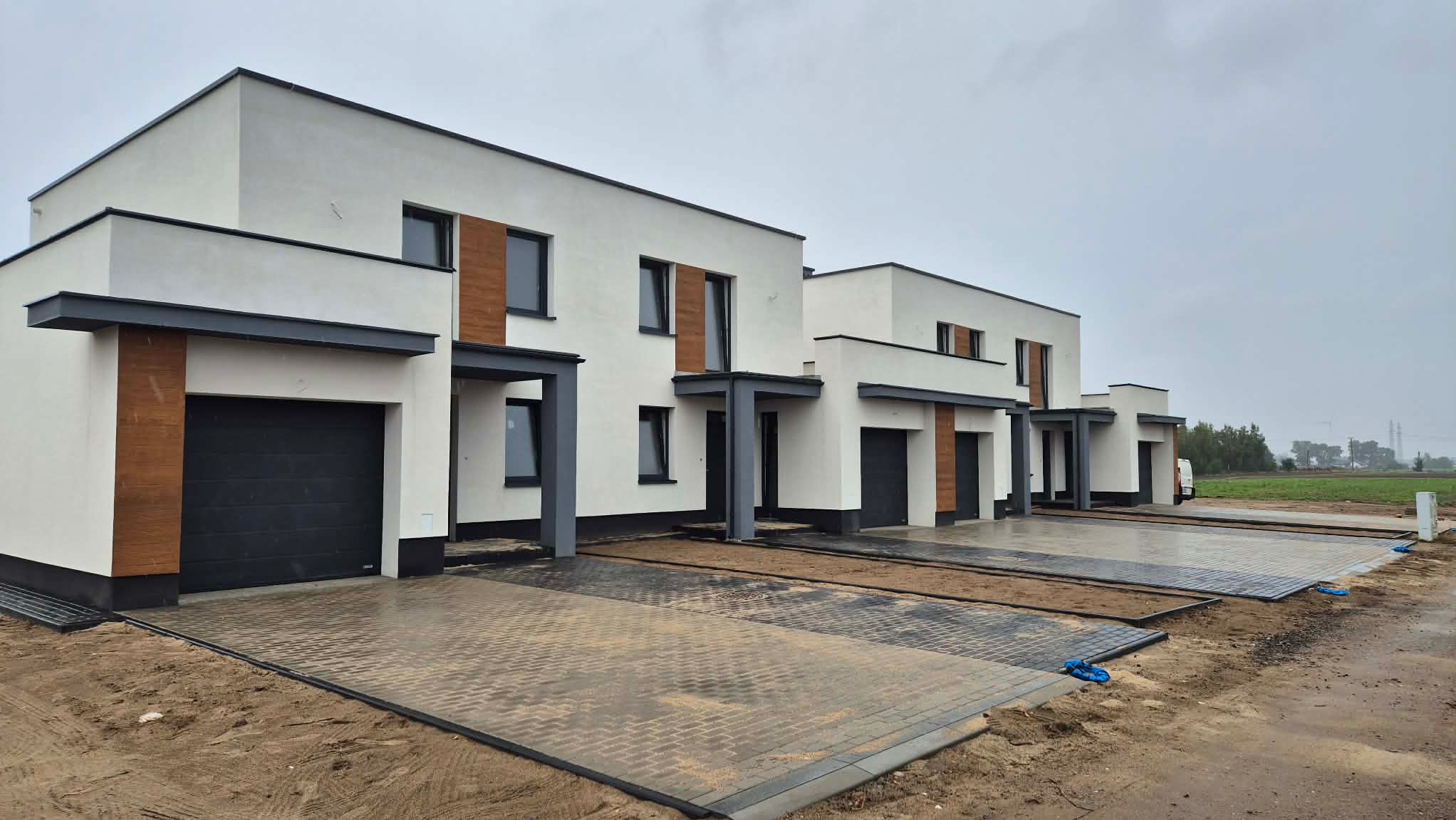 Nowoczesne, szeregowe domy z białymi elewacjami, grafitowymi akcentami i drewnianymi panelami. Kostka brukowa przed budynkami. Pochmurne niebo w tle.