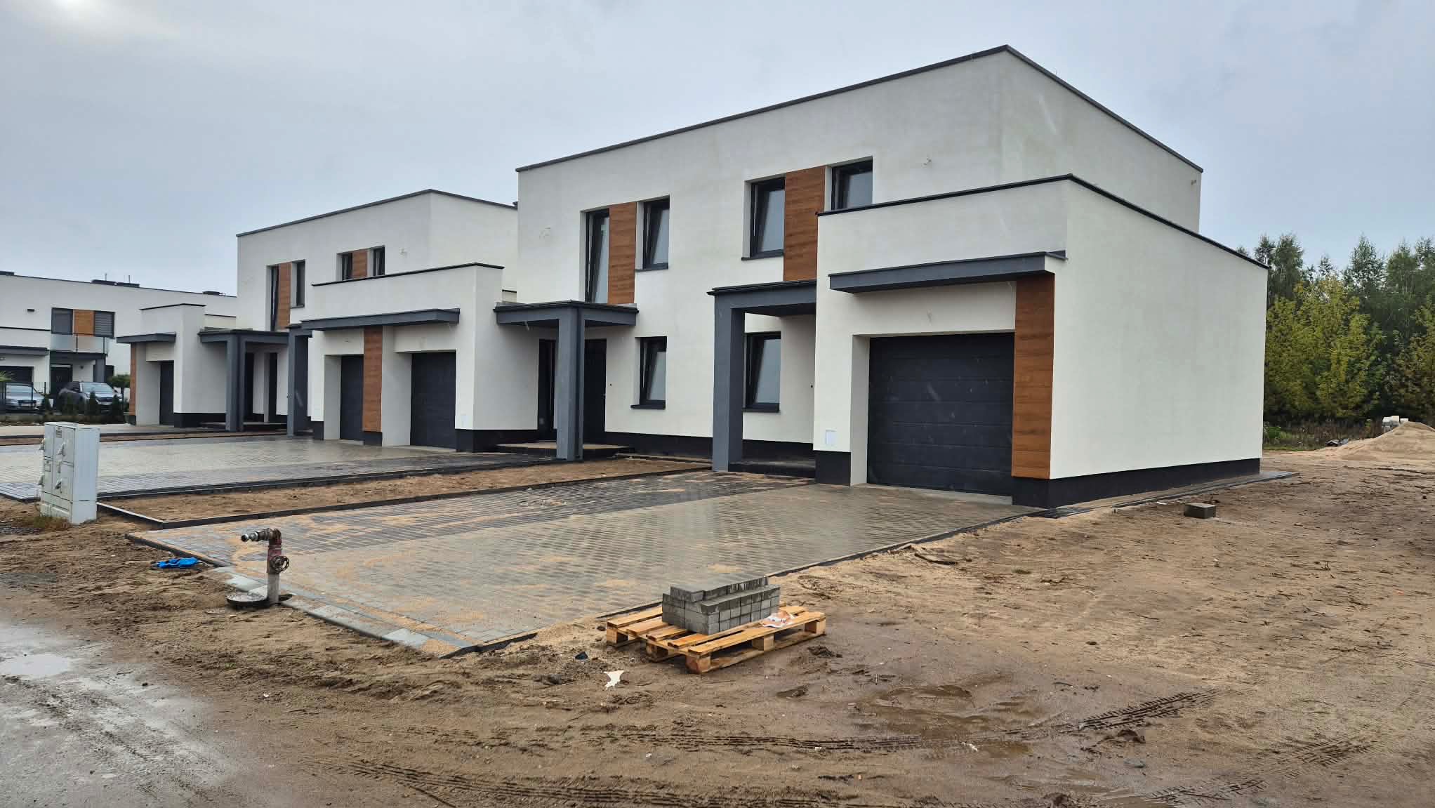 Nowoczesne, białe domy szeregowe z elementami drewna i szarą stolarką, świeżo po budowie, z kostką brukową na podjazdach i paletą kostki brukowej na pierwszym planie.
