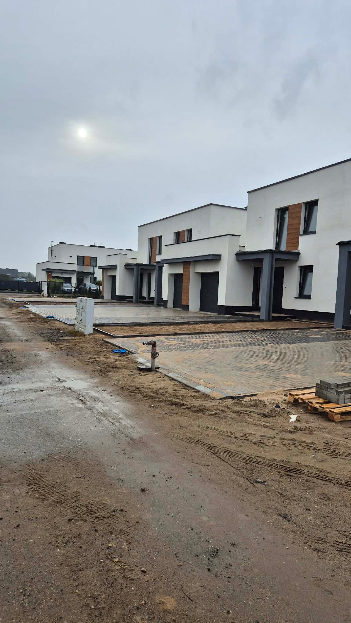 Nowoczesne, szeregowe domy z białymi elewacjami i elementami drewnopodobnymi, szara kostka brukowa na podjazdach, pochmurne niebo. Widok z poziomu gruntu.