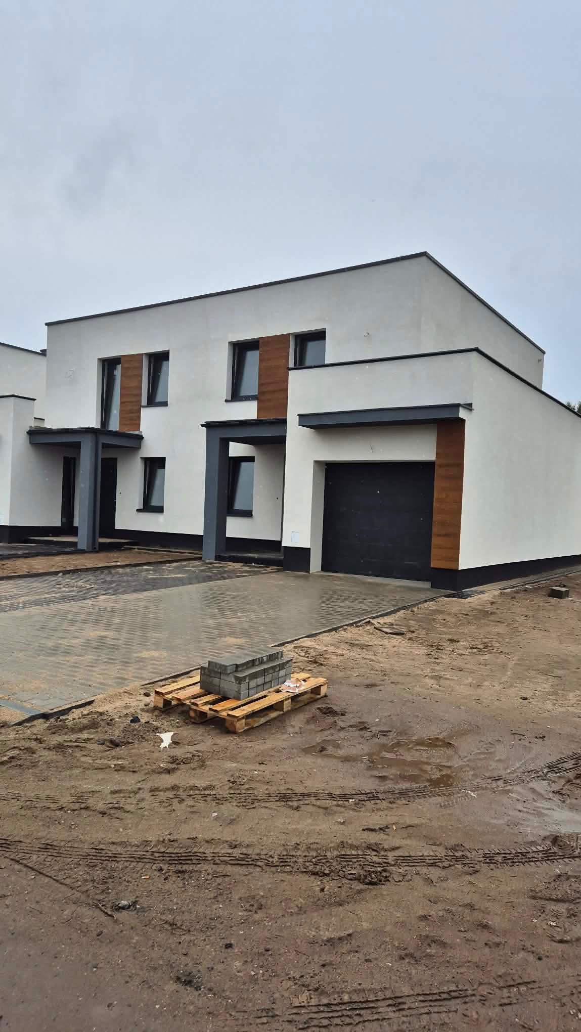 Nowoczesny dom z białą elewacją i drewnianymi akcentami, czarnymi ramami okiennymi i garażem. Na pierwszym planie paleta z kostką brukową.