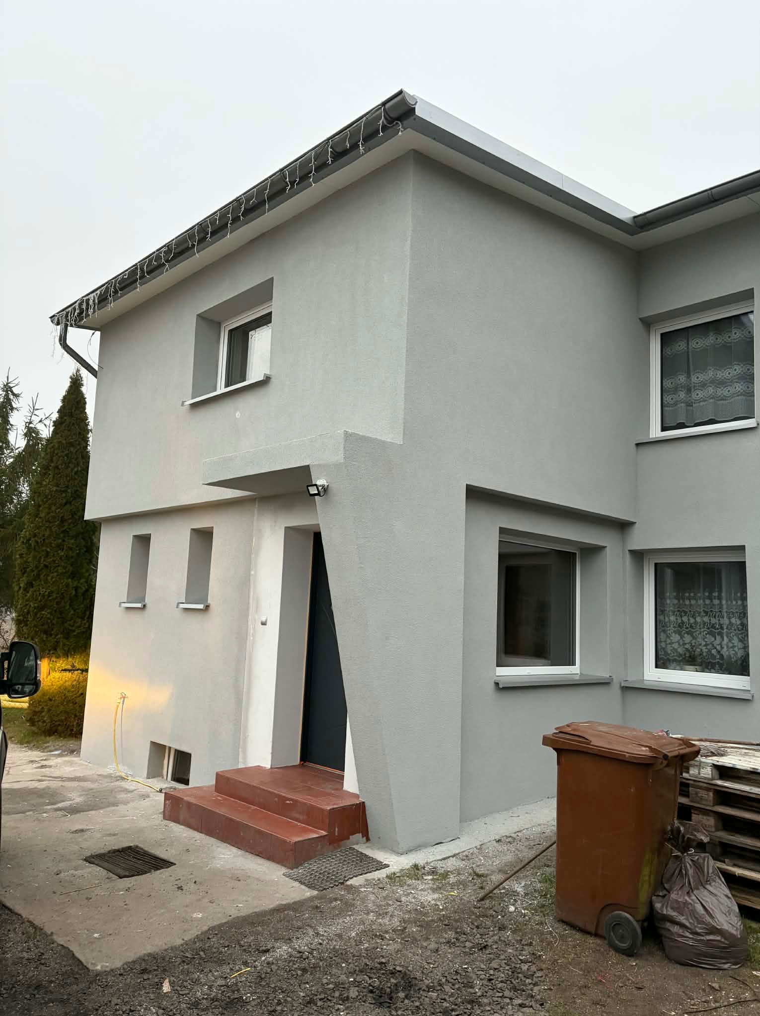 Dwukondygnacyjny dom z szarą elewacją, widoczne okna z białymi ramami, brązowe schody wejściowe i kosz na śmieci. Minimalistyczny styl, oświetlenie zewnętrzne.