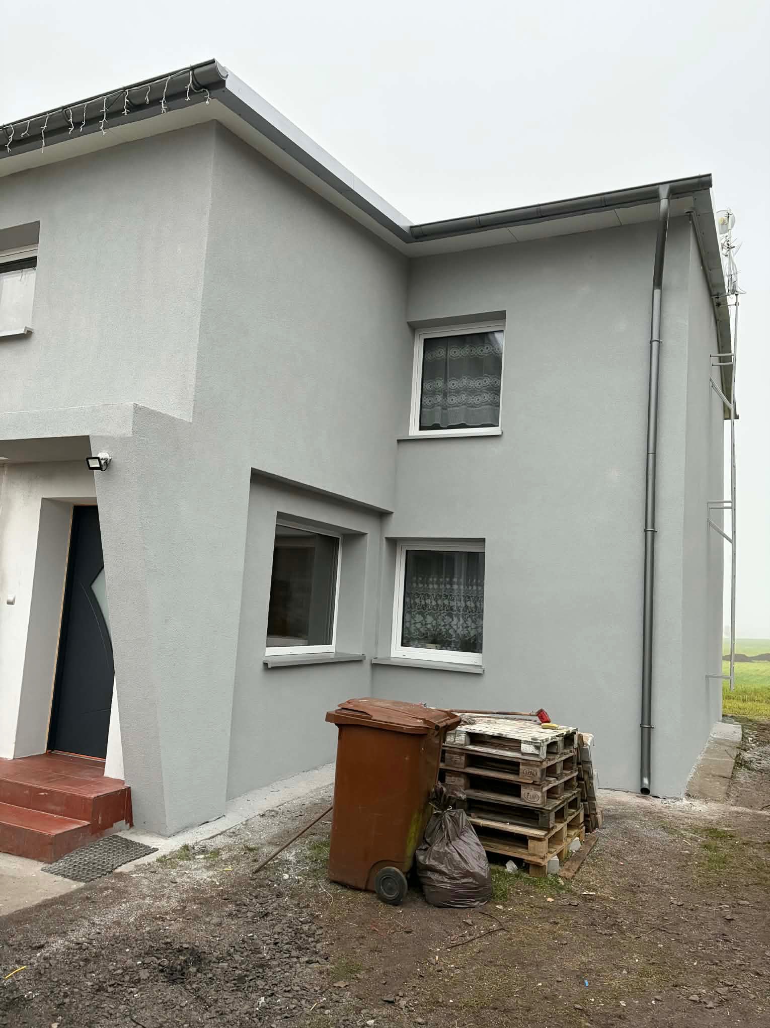 Dwukondygnacyjny dom z szarą elewacją po tynkowaniu, widoczne okna z firankami, rynna, palety i kosz na śmieci. Świeżo wykonane tynki elewacyjne.
