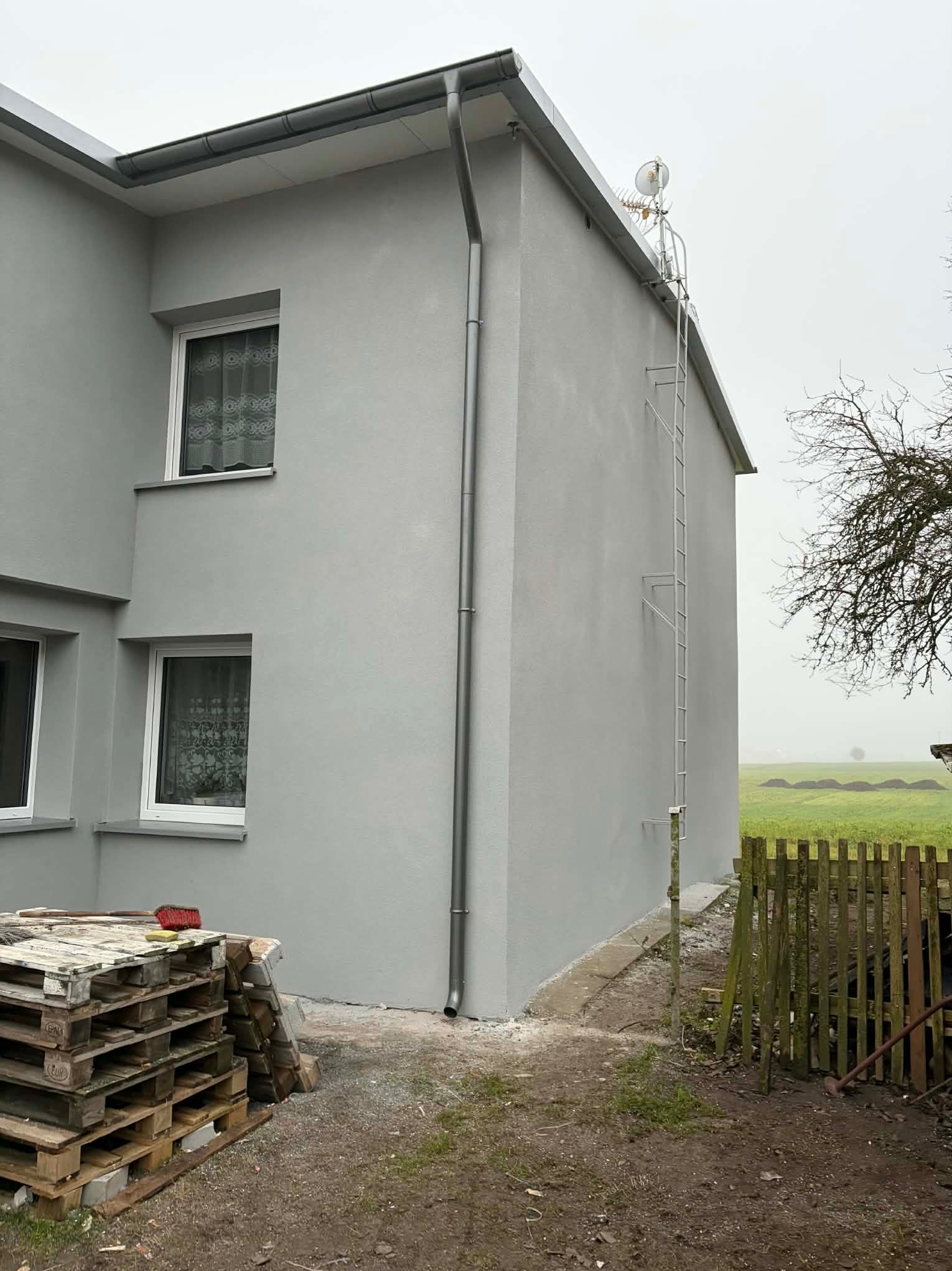 Szara elewacja domu jednorodzinnego z zamontowaną drabiną i anteną na dachu. Widoczne palety i fragment ogrodzenia. Ujęcie z dołu, pochmurny dzień.
