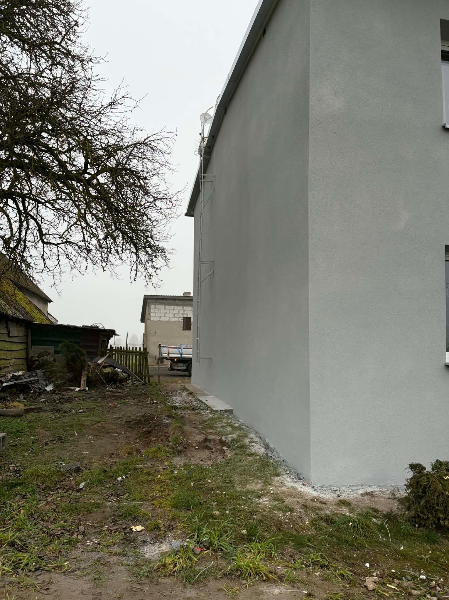 Świeżo otynkowana, szara elewacja domu jednorodzinnego z drabinką, widoczna z boku na tle pochmurnego nieba i zabudowań gospodarczych.