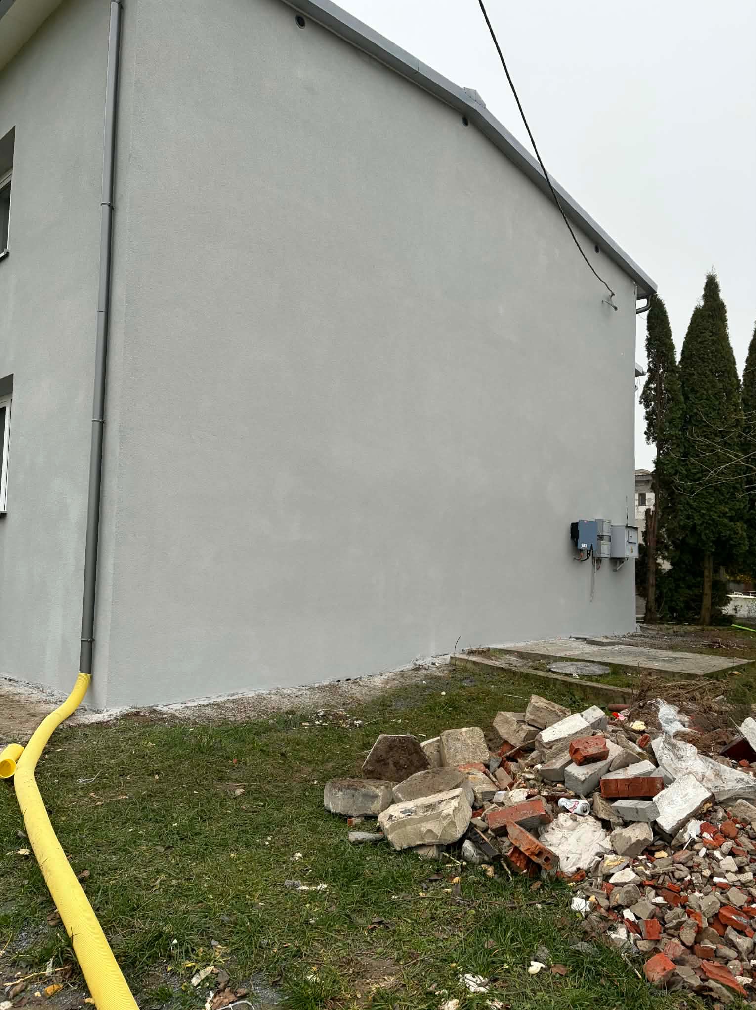 Jasnoszara elewacja budynku z rynną i skrzynkami elektrycznymi. Na trawniku gruz i żółty wąż. W tle wysokie drzewa iglaste.