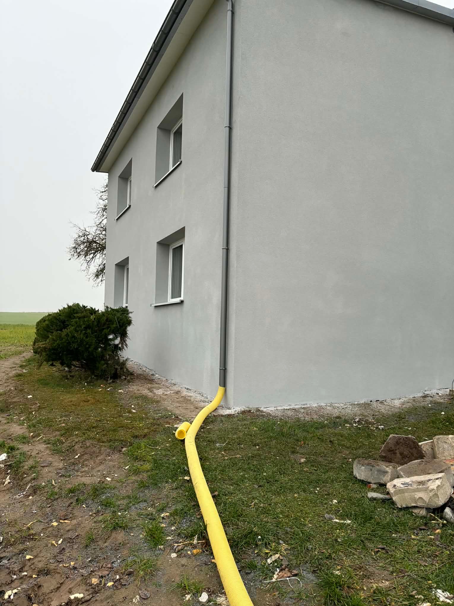 Świeżo otynkowana, szara elewacja domu jednorodzinnego z widoczną rurą spustową i żółtym wężem na trawniku. Minimalistyczny styl, pochmurny dzień.