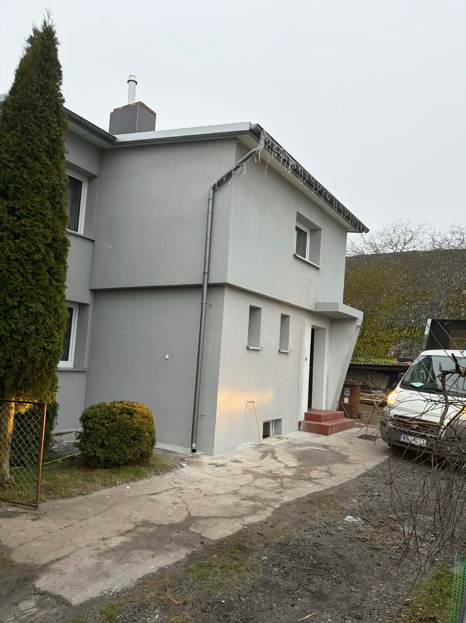 Dwukondygnacyjny dom po tynkowaniu elewacji na szaro, z widocznymi detalami wykończeniowymi, rynną i fragmentem podjazdu. Ujęcie z dołu, pochmurne niebo.