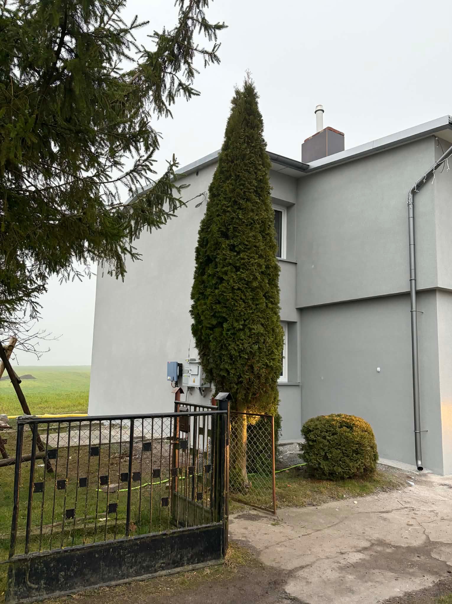 Szary dom jednorodzinny z nową elewacją, widoczny zza metalowego ogrodzenia i zieleni. Świeżo otynkowana ściana, rynna i komin. Mglista pogoda.