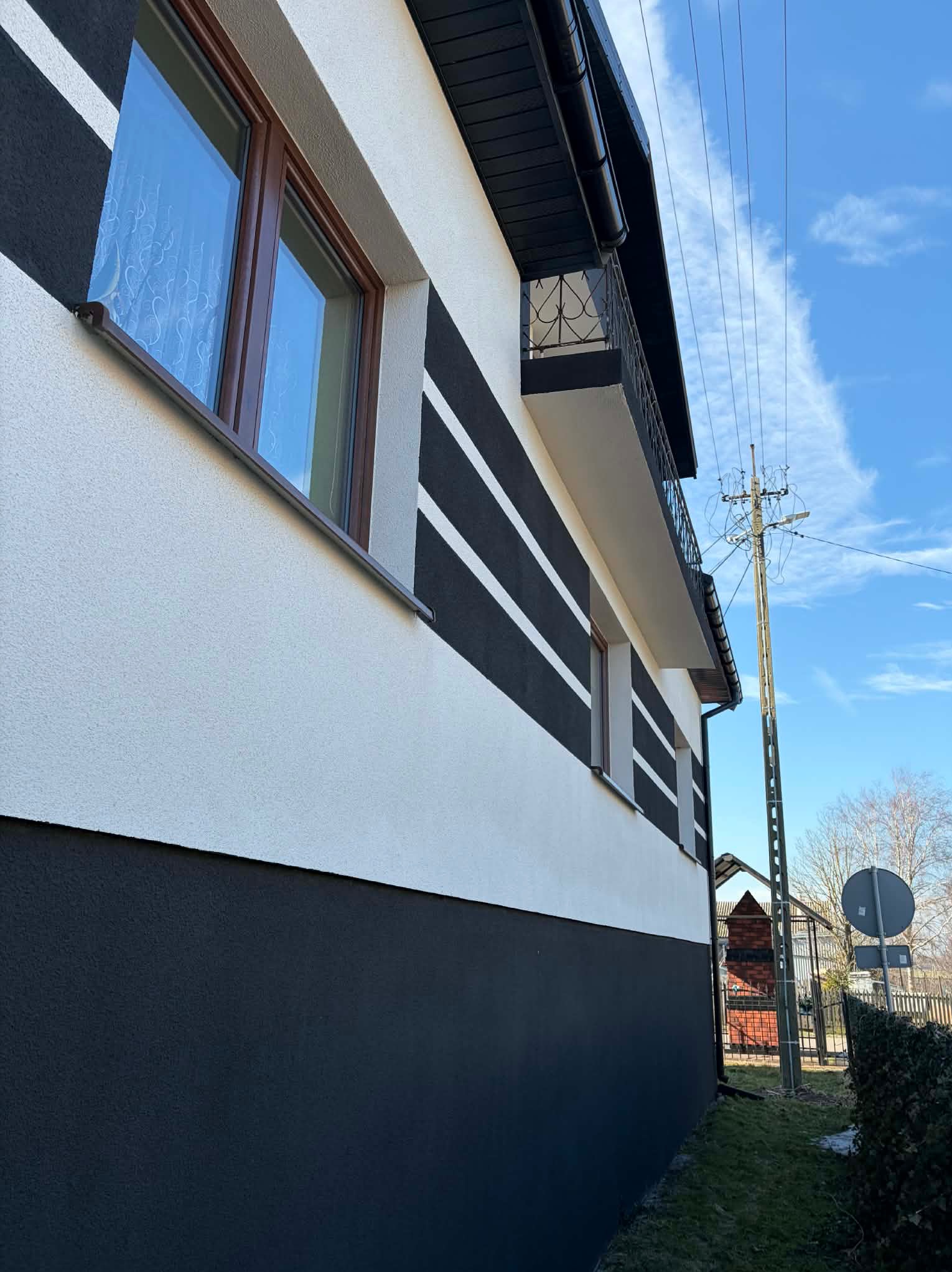 Elewacja domu po malowaniu w biało-czarne pasy, z widocznym balkonem i oknami z brązowymi ramami. Słup elektryczny w tle.