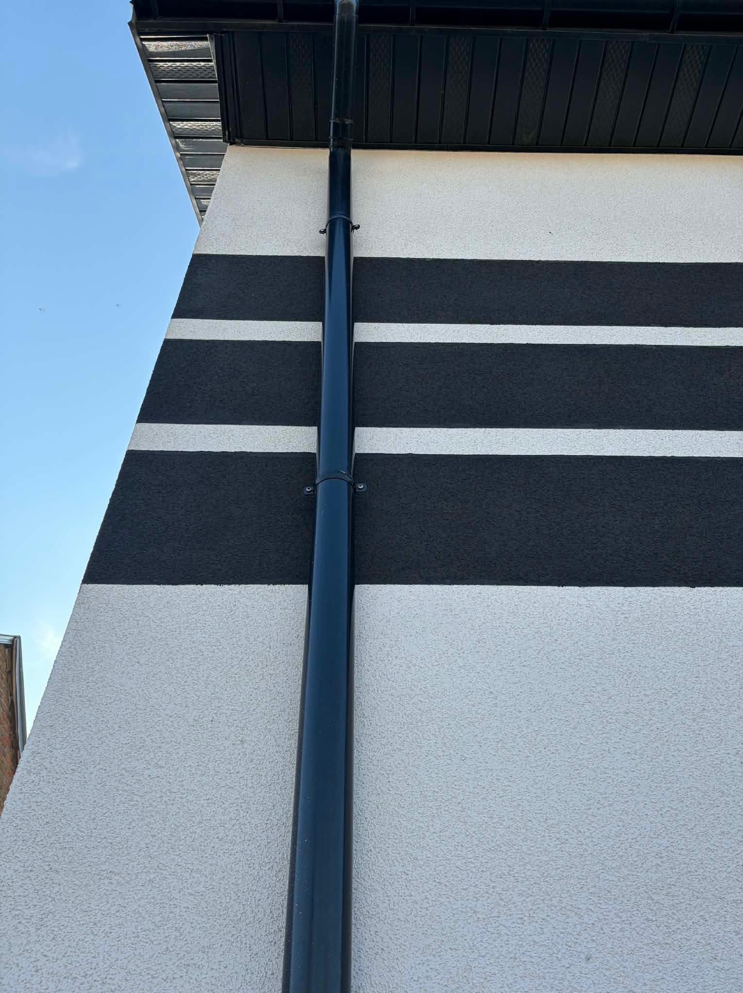 Elewacja budynku z dekoracyjnymi, poziomymi pasami w kolorze grafitowym i jasnoszarym, z widoczną rynną. Tekstura tynku barankowego.