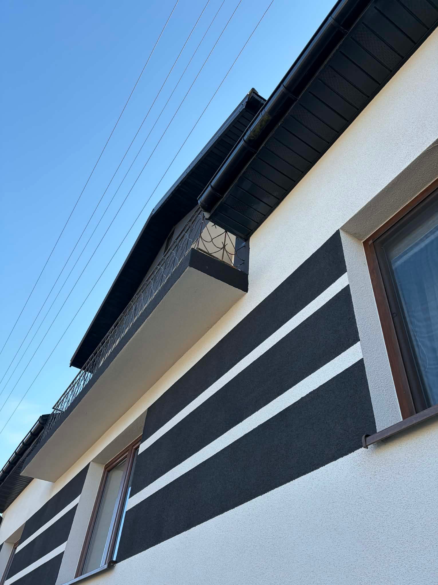 Elewacja domu z białym tynkiem i czarnymi pasami, widok z dołu, okna z brązowymi ramami, balkon z czarną balustradą, błękitne niebo w tle.