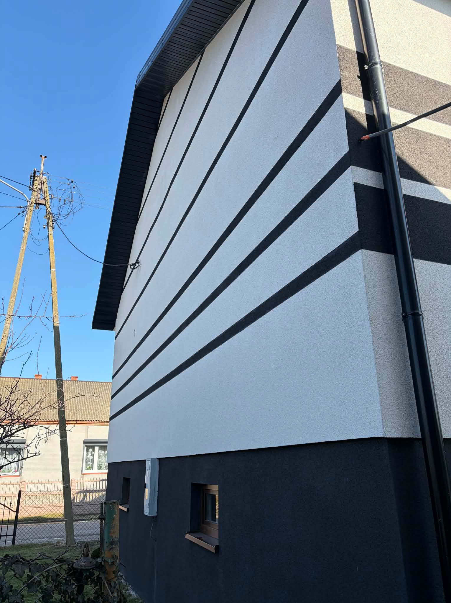 Boczny widok domu po renowacji elewacji: biała ściana z poziomymi czarnymi pasami, ciemny cokół i rynna. Widoczne okna i słup elektryczny w tle.