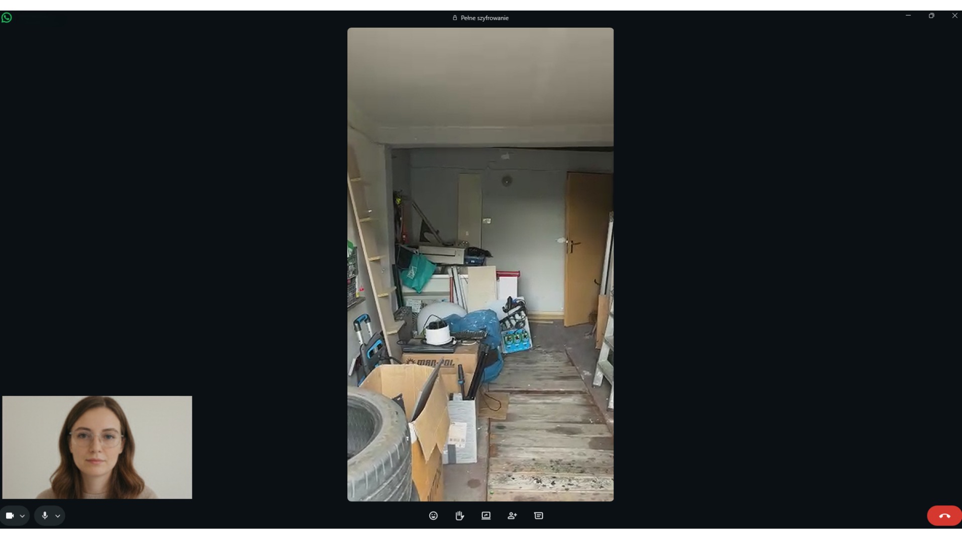 Ekran rozmowy wideo z widokiem zagraconego garażu: narzędzia, opony, kartony. Widoczna drabina i drzwi. U dołu okno z twarzą kobiety.