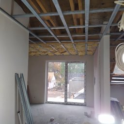 Serhii Moiseiev Renonty Poznań i Okolice - Wnętrze w trakcie prac wykończeniowych: widoczna konstrukcja sufitu podwieszanego z izolacją, okno z widokiem na zewnątrz i materiały budowlane na podłodze.