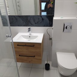 Serhii Moiseiev Renonty Poznań i Okolice - Nowoczesna łazienka po remoncie: białe płytki, drewniana szafka pod umywalką, toaleta podwieszana. W lustrze odbija się osoba robiąca zdjęcie.
