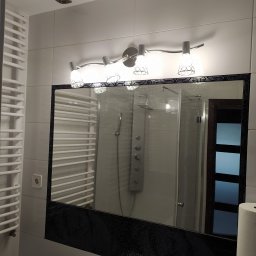 Serhii Moiseiev Renonty Poznań i Okolice - Nowoczesna łazienka z białymi płytkami, grzejnikiem drabinkowym i lustrem z mozaikową ramą. Widoczna armatura i oświetlenie.