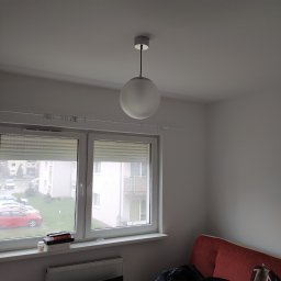Serhii Moiseiev Renonty Poznań i Okolice - Wnętrze pokoju z oknem i kulistą lampą sufitową. Widoczne rolety okienne, grzejnik pod oknem, sofa z ubraniami i plecakiem. Świeżo pomalowane ściany.