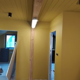 Serhii Moiseiev Renonty Poznań i Okolice - Oliwkowa ściana z drewnianą belką i oświetleniem LED. Widoczne przygotowania do dalszych prac wykończeniowych, zabezpieczona podłoga i otwarte przejście do innego pomieszczenia.