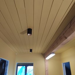 Serhii Moiseiev Renonty Poznań i Okolice - Żółte ściany i sufit z drewnianą belką oraz dwoma czarnymi lampami punktowymi. Widoczne otwory drzwiowe i okno zabezpieczone folią malarską.