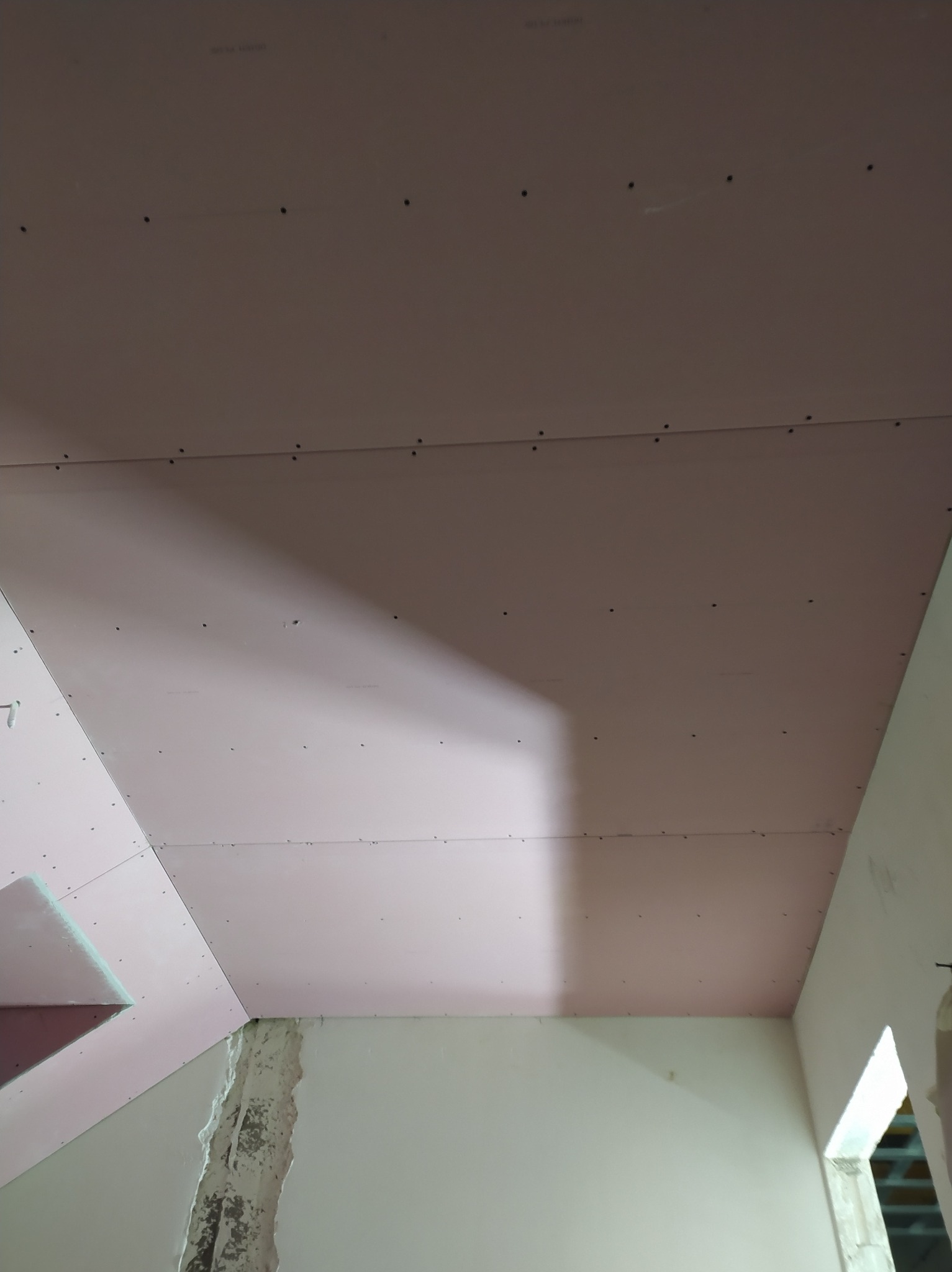 Różowe płyty gipsowo-kartonowe zamontowane na suficie, widoczne ślady po wkrętach, fragment ściany z widocznymi uszkodzeniami. Prace wykończeniowe w trakcie.