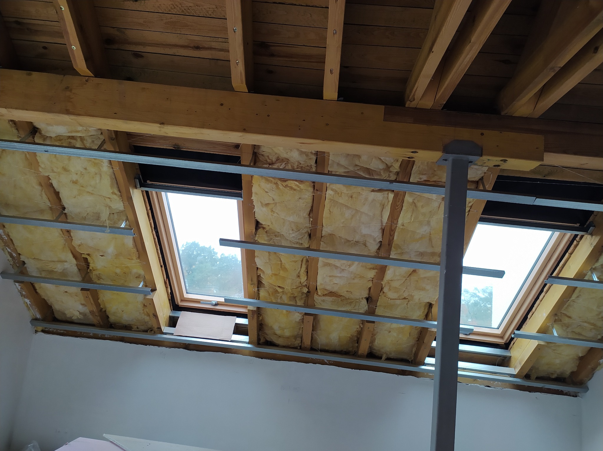 Montaż izolacji z wełny mineralnej na poddaszu z oknami dachowymi. Widoczne drewniane belki, metalowe profile i fragment białej ściany. Prace wykończeniowe.