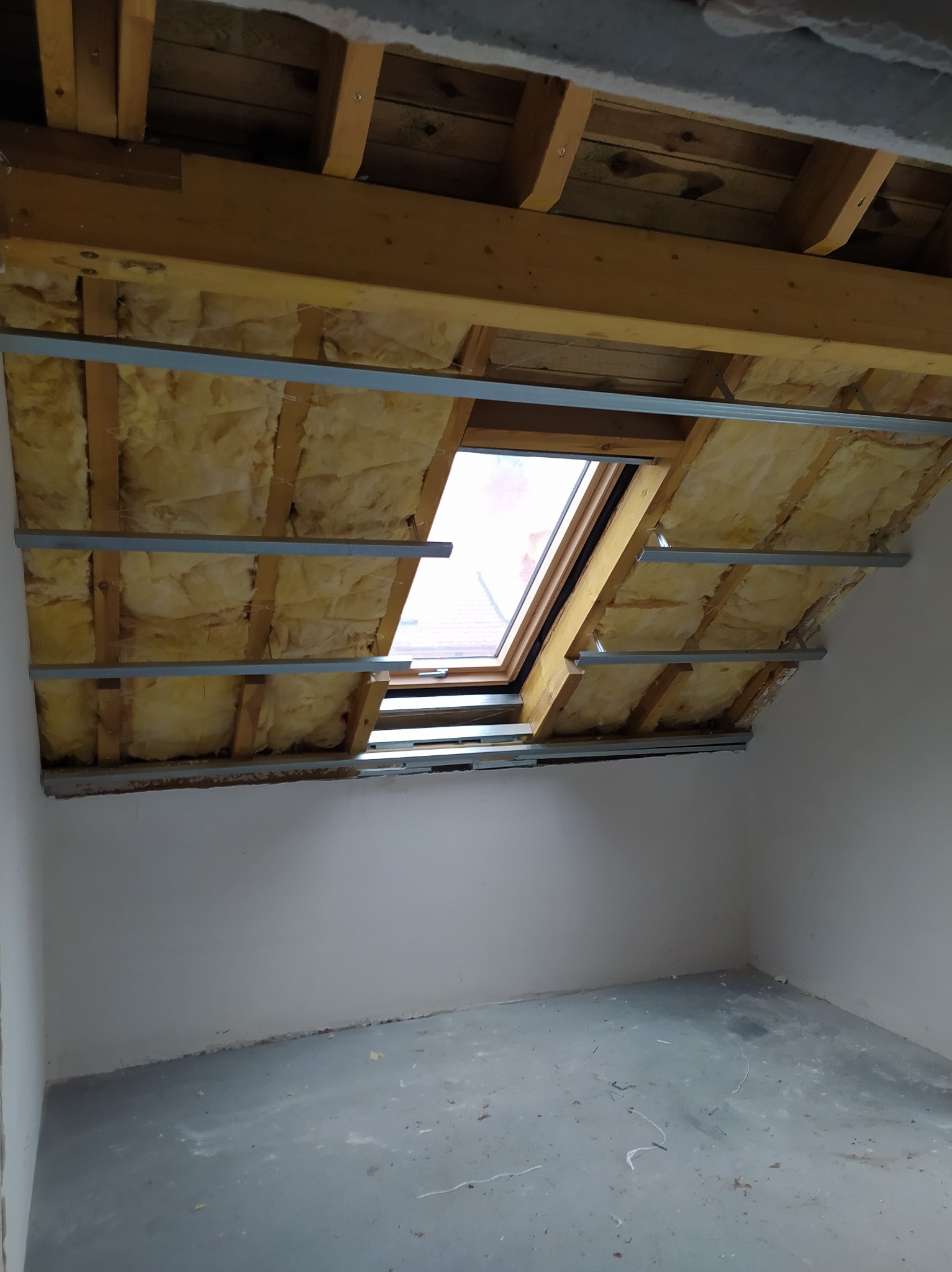 Izolacja poddasza z oknem dachowym, widoczne drewniane belki, wełna mineralna i metalowe profile. Ściany w surowym stanie, przygotowane do dalszych prac wykończeniowych.