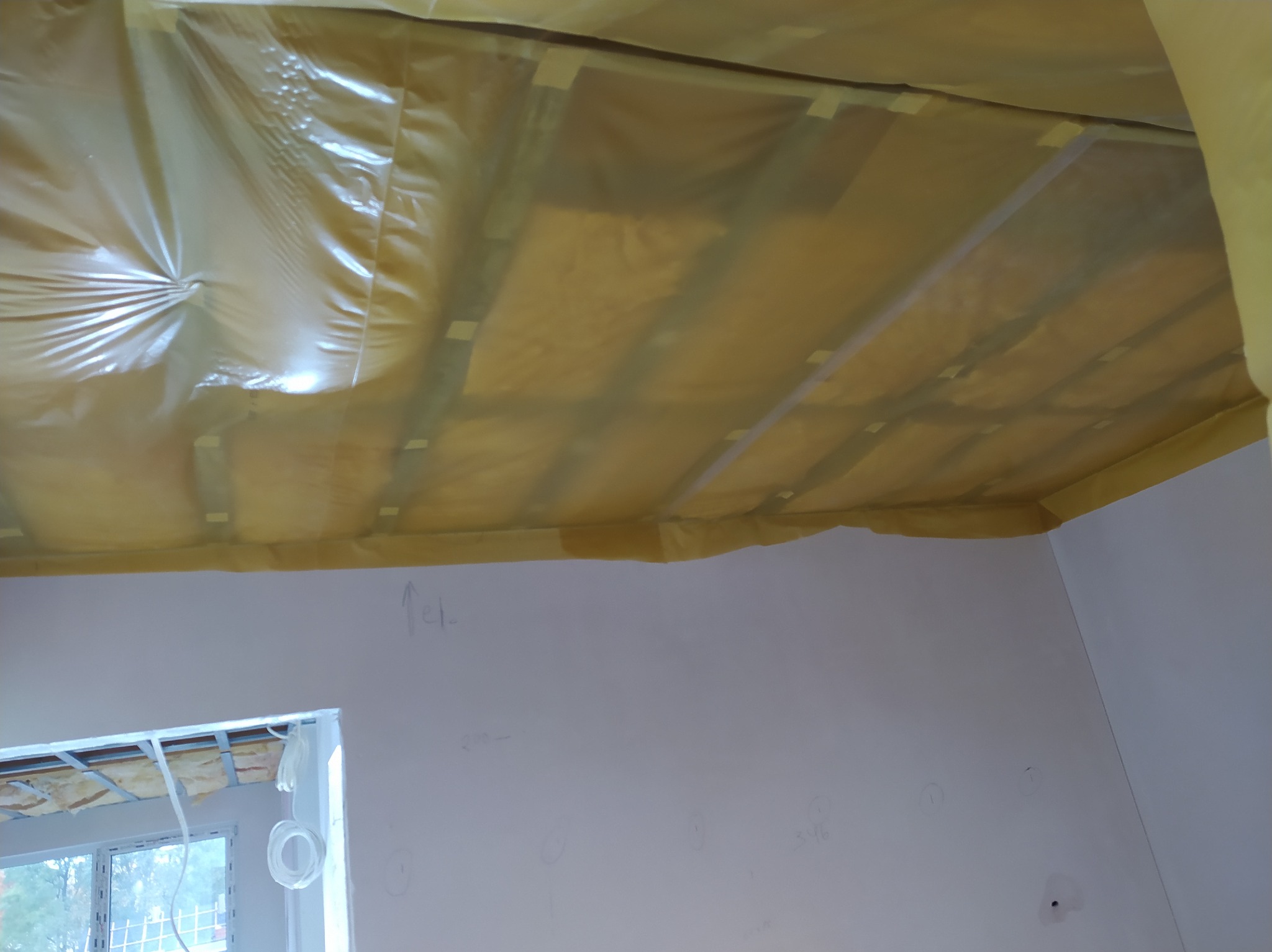 Izolacja poddasza z folią paroizolacyjną, zabezpieczona taśmą. Widoczne fragmenty okna i ściany z notatkami. Prace remontowe w trakcie realizacji.