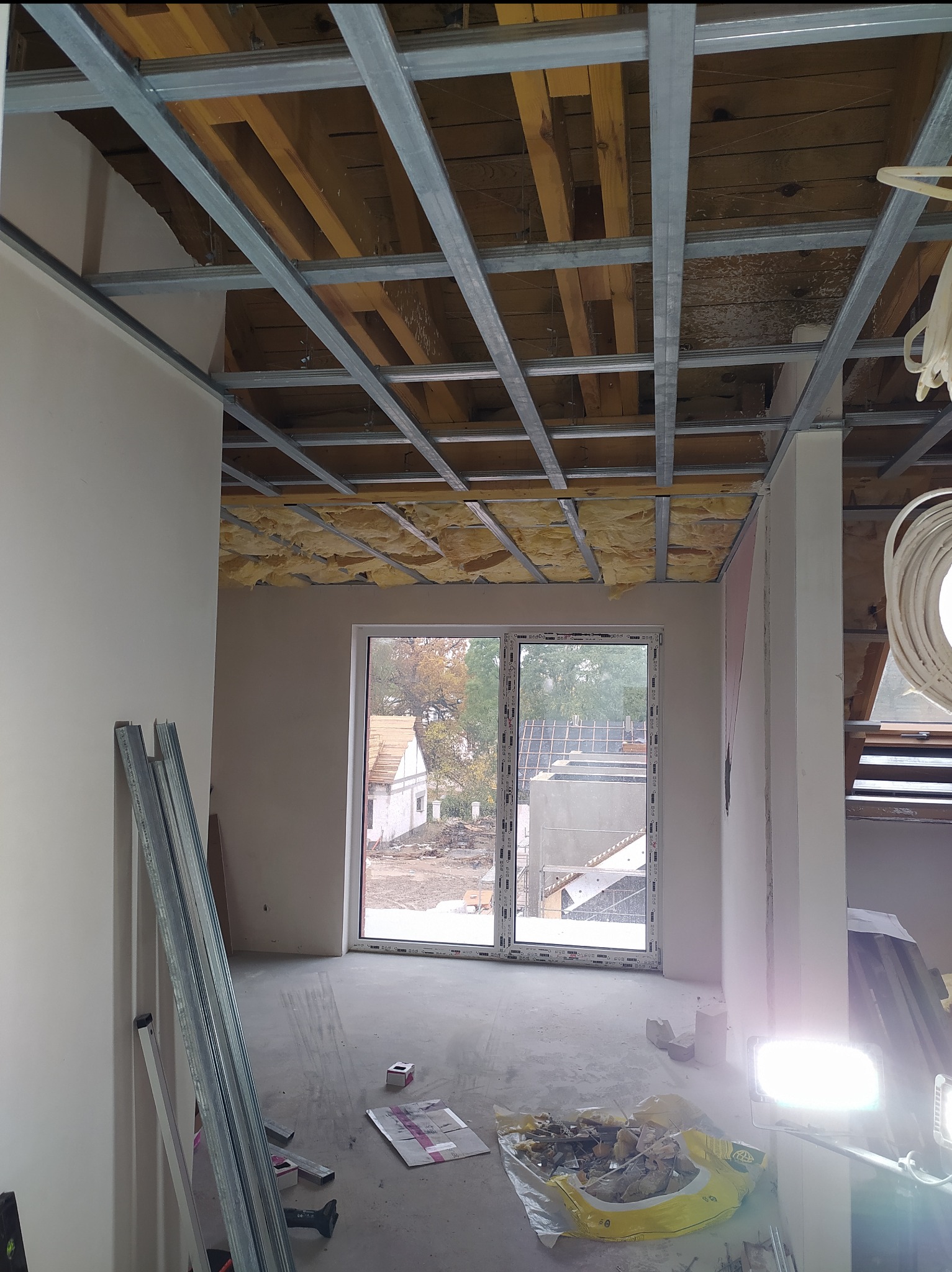 Wnętrze w trakcie prac wykończeniowych: widoczna konstrukcja sufitu podwieszanego z izolacją, okno z widokiem na zewnątrz i materiały budowlane na podłodze.