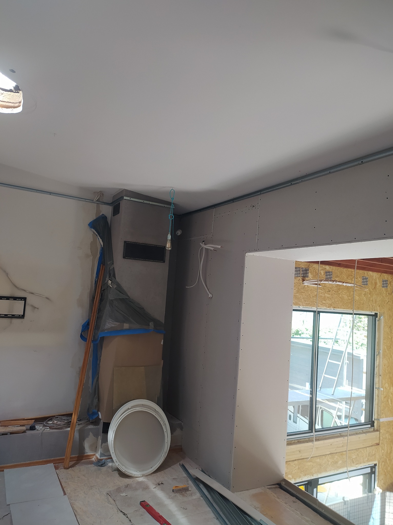 Wnętrze w trakcie prac wykończeniowych: zabudowa kominka zabezpieczona folią, ściany z płyt karton-gips, widoczne przewody elektryczne i narzędzia. Okno z widokiem na zewnątrz.