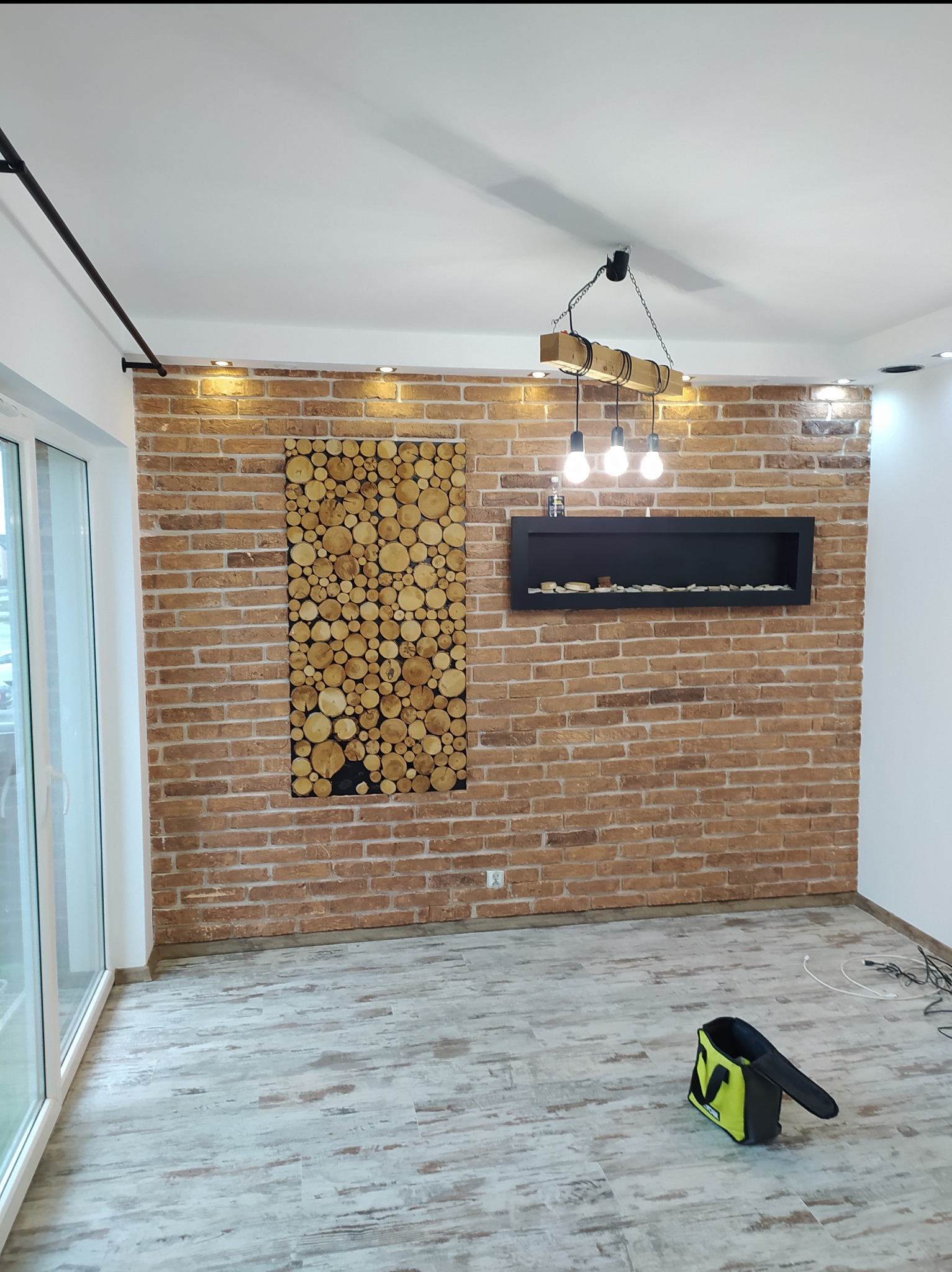 Wykończona ściana z cegły z dekoracyjnym panelem z drewna i czarnym wnękowym elementem. Podłoga wyłożona panelami imitującymi drewno. Widoczna torba narzędziowa.