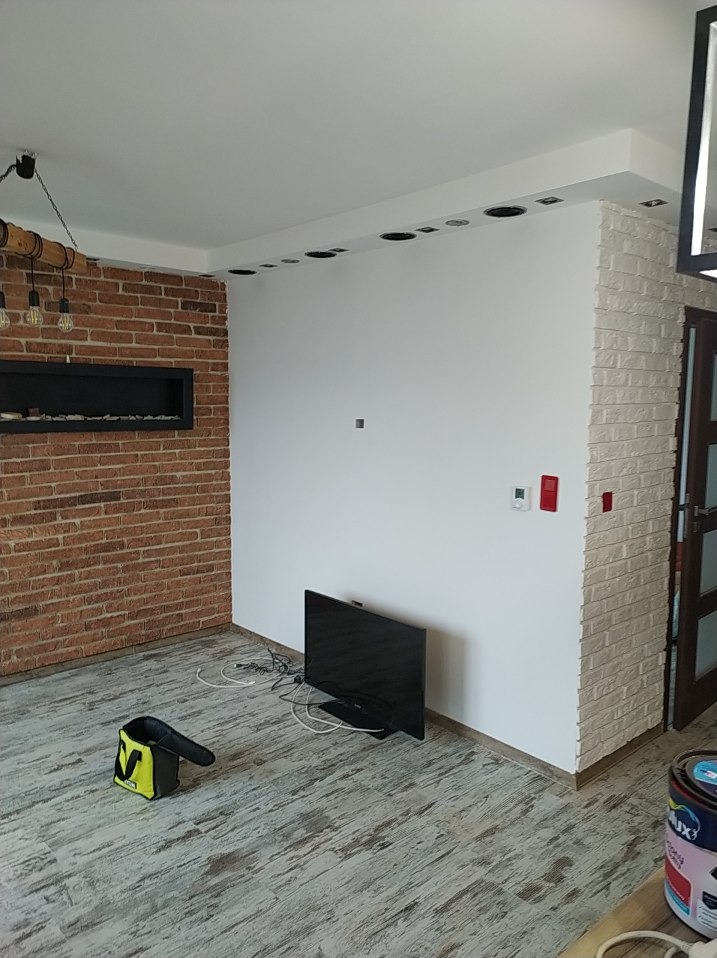 Wnętrze pokoju po remoncie: ściana z cegły, biała ściana z otworami na oświetlenie, imitacja cegły na rogu, podłoga imitująca drewno, telewizor i torba narzędziowa.