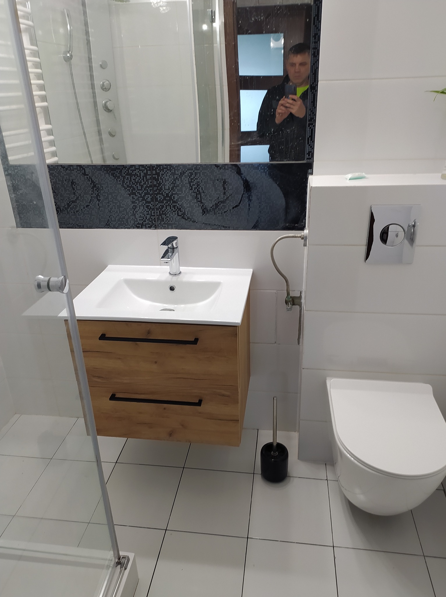 Nowoczesna łazienka po remoncie: białe płytki, drewniana szafka pod umywalką, toaleta podwieszana. W lustrze odbija się osoba robiąca zdjęcie.