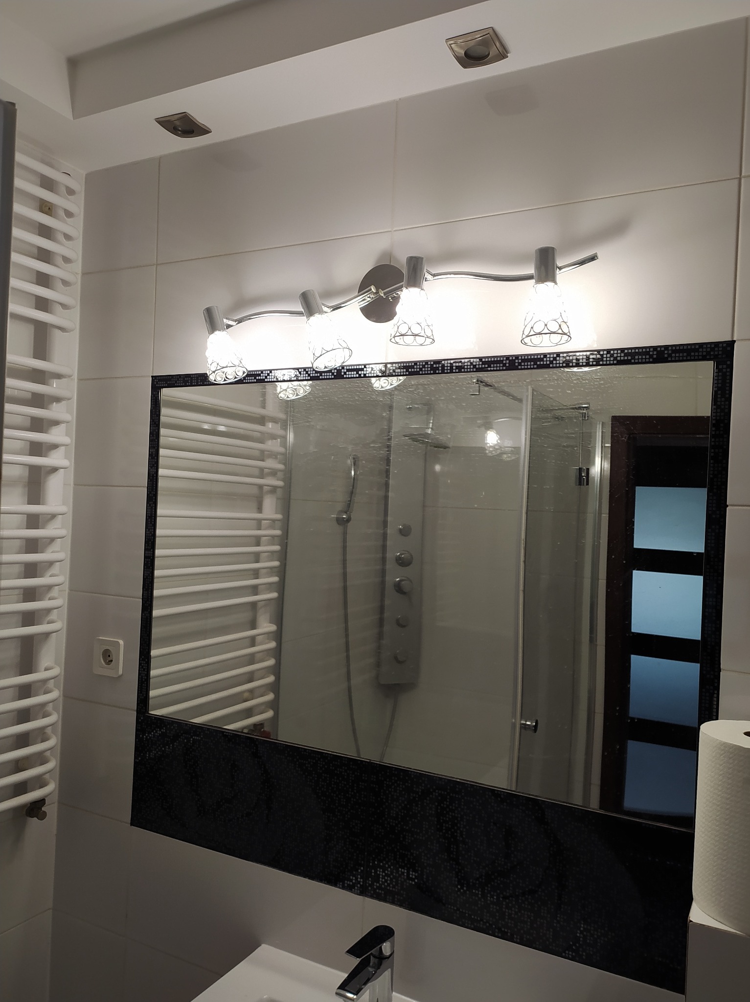 Nowoczesna łazienka z białymi płytkami, grzejnikiem drabinkowym i lustrem z mozaikową ramą. Widoczna armatura i oświetlenie.