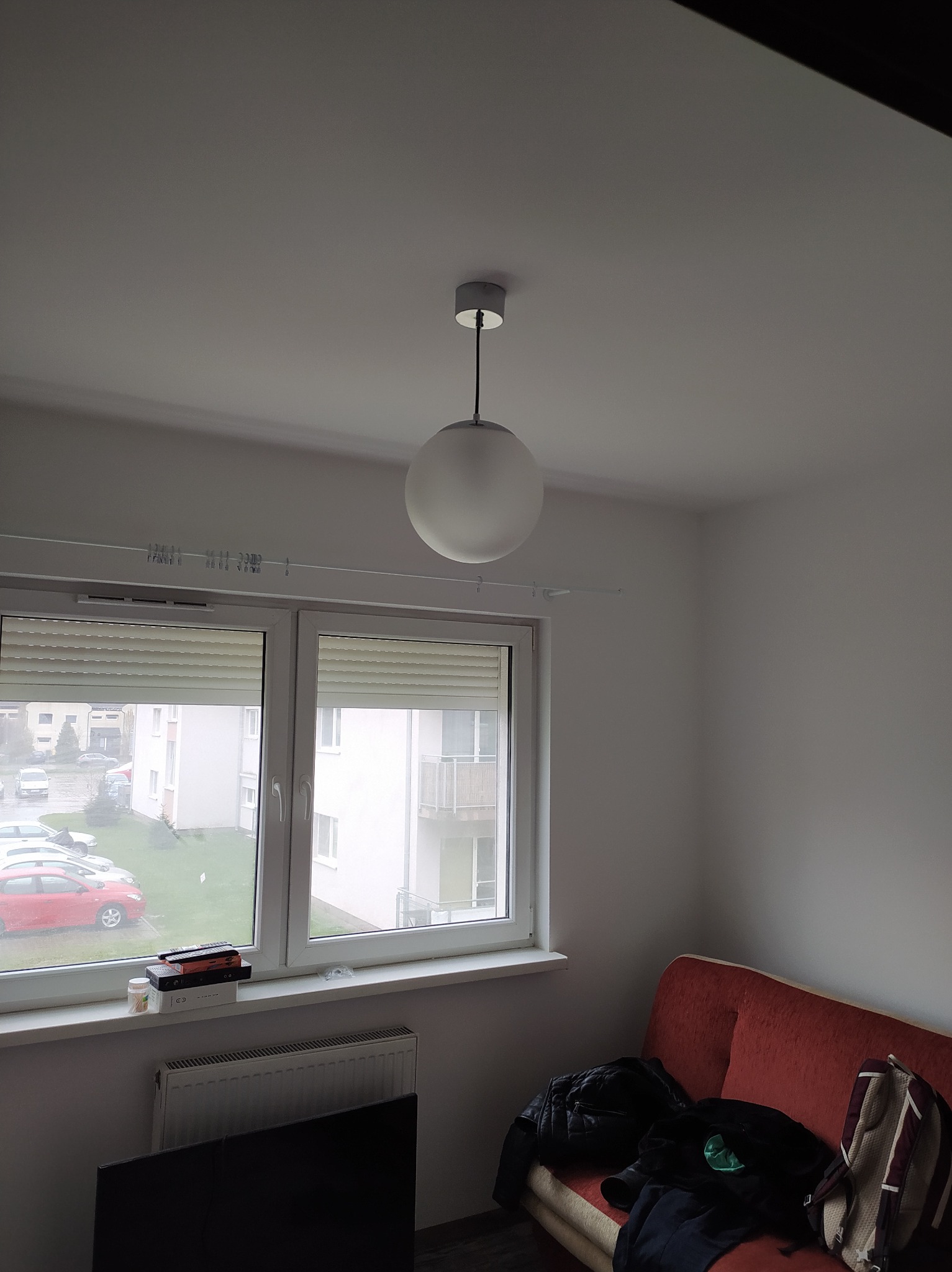 Wnętrze pokoju z oknem i kulistą lampą sufitową. Widoczne rolety okienne, grzejnik pod oknem, sofa z ubraniami i plecakiem. Świeżo pomalowane ściany.