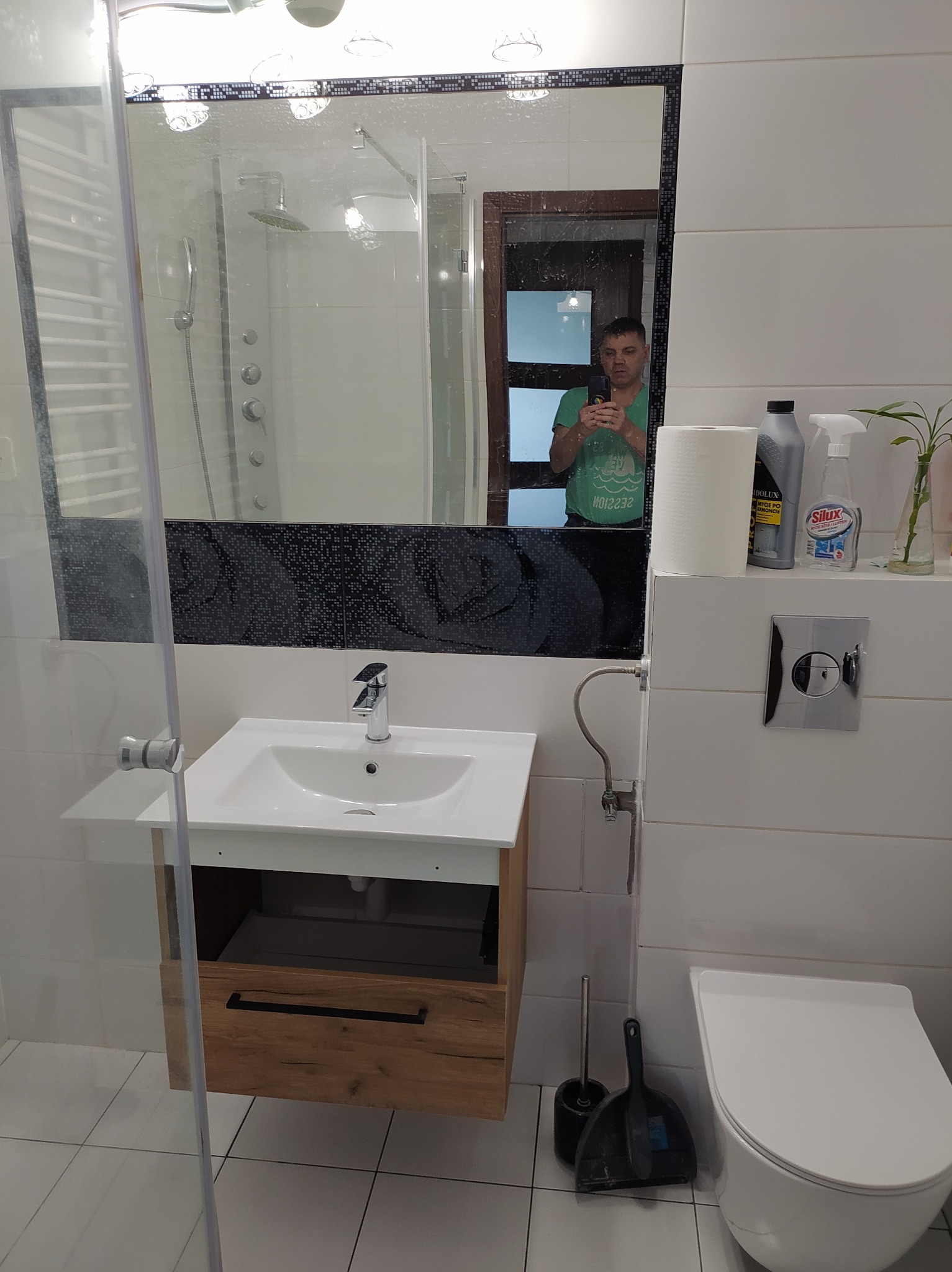 Nowoczesna łazienka z kabiną prysznicową, umywalką z szafką z drewnianym frontem i toaletą. W lustrze odbija się osoba robiąca zdjęcie.