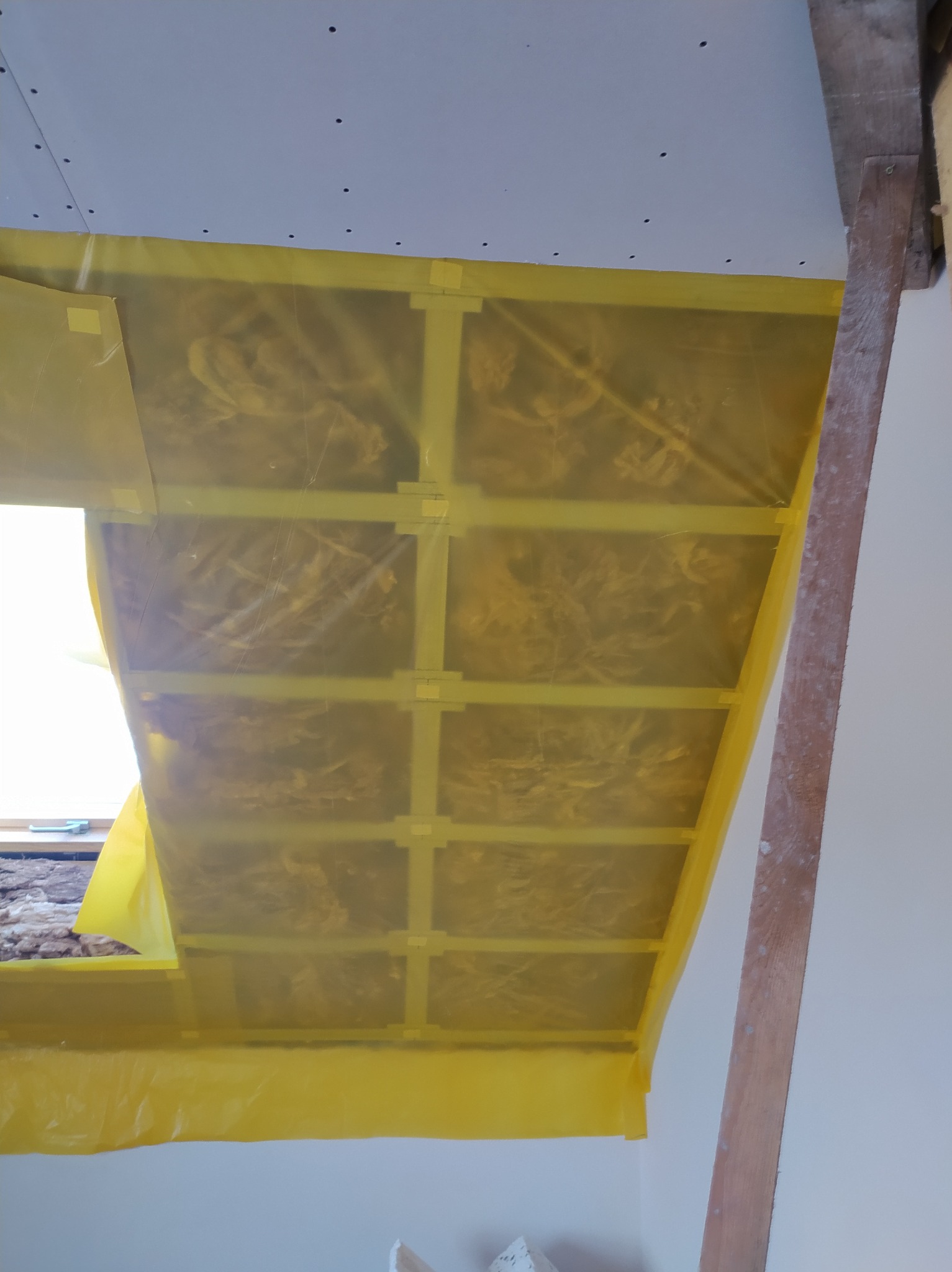 Ocieplenie poddasza z widoczną folią paroizolacyjną, zabezpieczającą wełnę mineralną. Widoczny fragment okna dachowego i drewniana konstrukcja więźby.