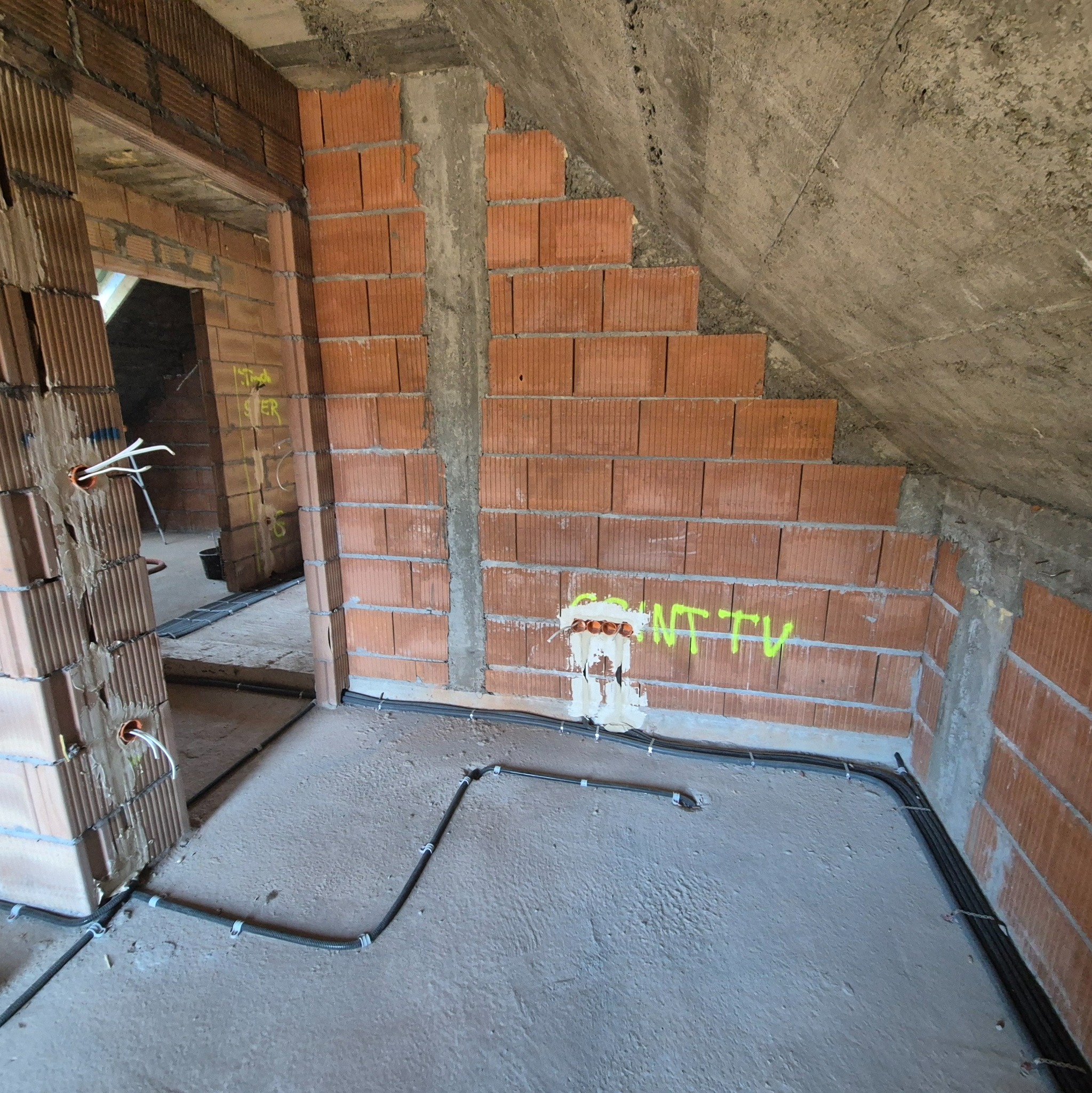 Instalacja elektryczna w surowym wnętrzu poddasza: kable w peszlach prowadzone po podłodze, puszki elektryczne w ścianach z cegły. Widoczne napisy na ścianie.