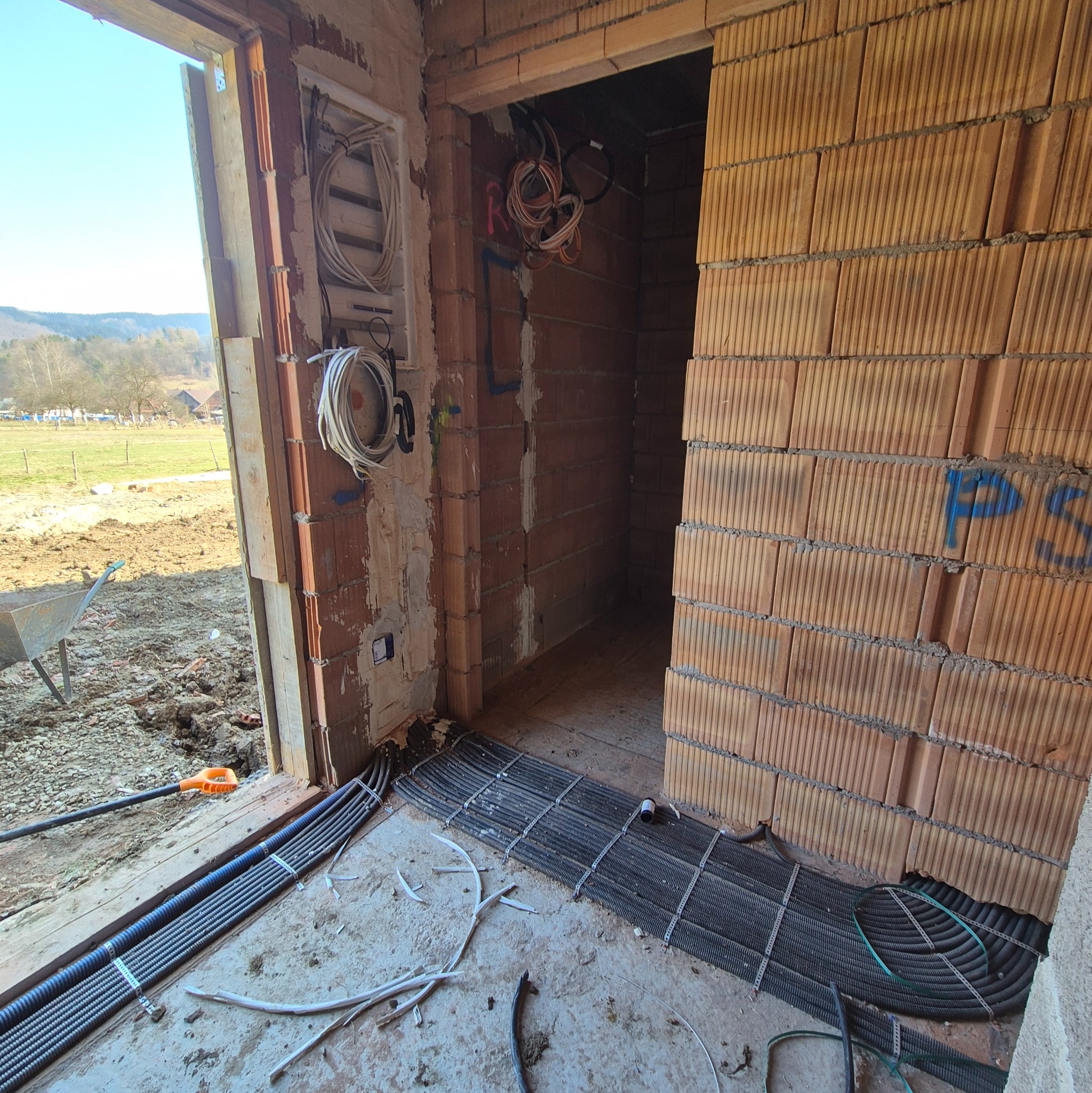 Wnętrze w trakcie budowy z widocznymi przewodami elektrycznymi ułożonymi na podłodze i zwisającymi ze ściany, ceglane ściany, surowy stan, okno z widokiem na zewnątrz.