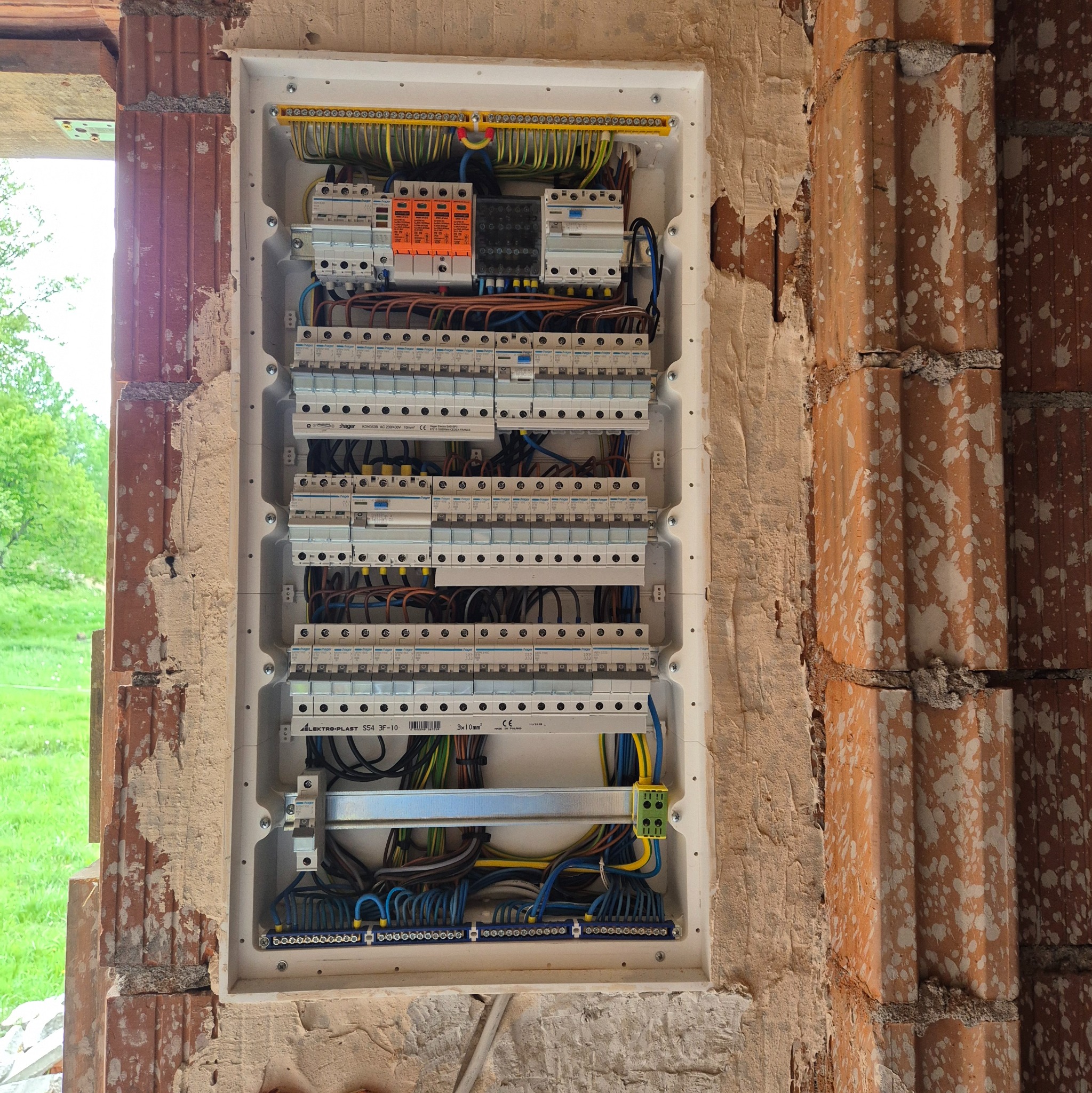 Rozdzielnica elektryczna w trakcie montażu, widoczne liczne przewody i zabezpieczenia, osadzona w surowej ścianie z cegły. Widok z bliska na wnętrze skrzynki.