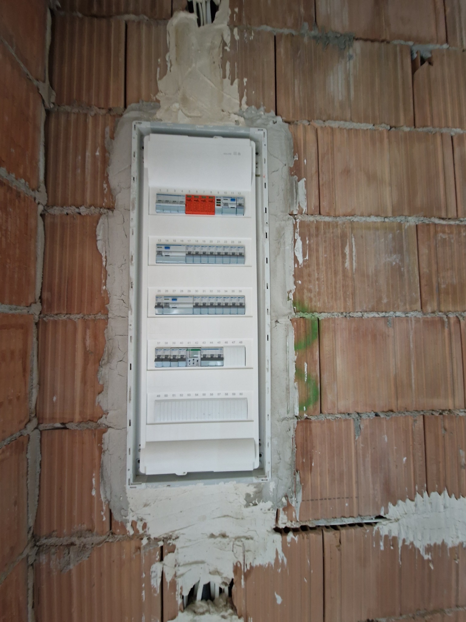 Biała skrzynka rozdzielcza z bezpiecznikami, zamontowana w surowej, ceglanej ścianie z widocznymi śladami zaprawy. Widok z bliska na etapie montażu.