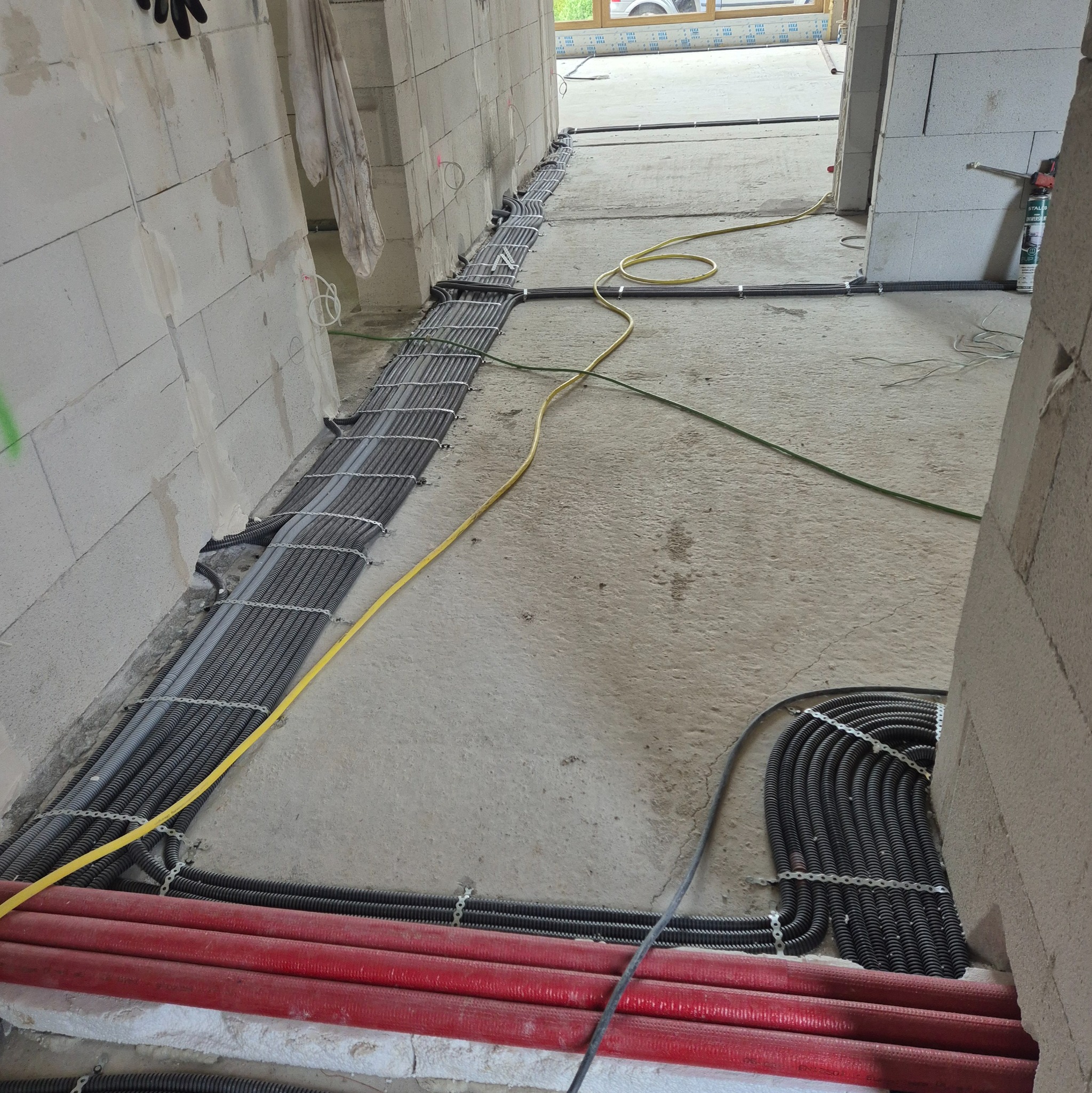 Instalacja przewodów elektrycznych w peszlach, mocowanych metalowymi uchwytami do surowej posadzki w trakcie budowy. Widoczne przewody w kolorach szarym, czerwonym i żółtym.