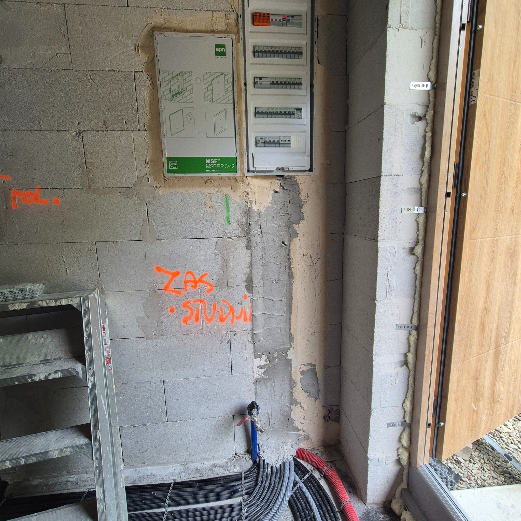 Wewnętrzna instalacja elektryczna w surowym stanie: rozdzielnia z bezpiecznikami na ścianie z betonu komórkowego, rury instalacyjne przy podłodze, widoczne oznaczenia na ścianie.