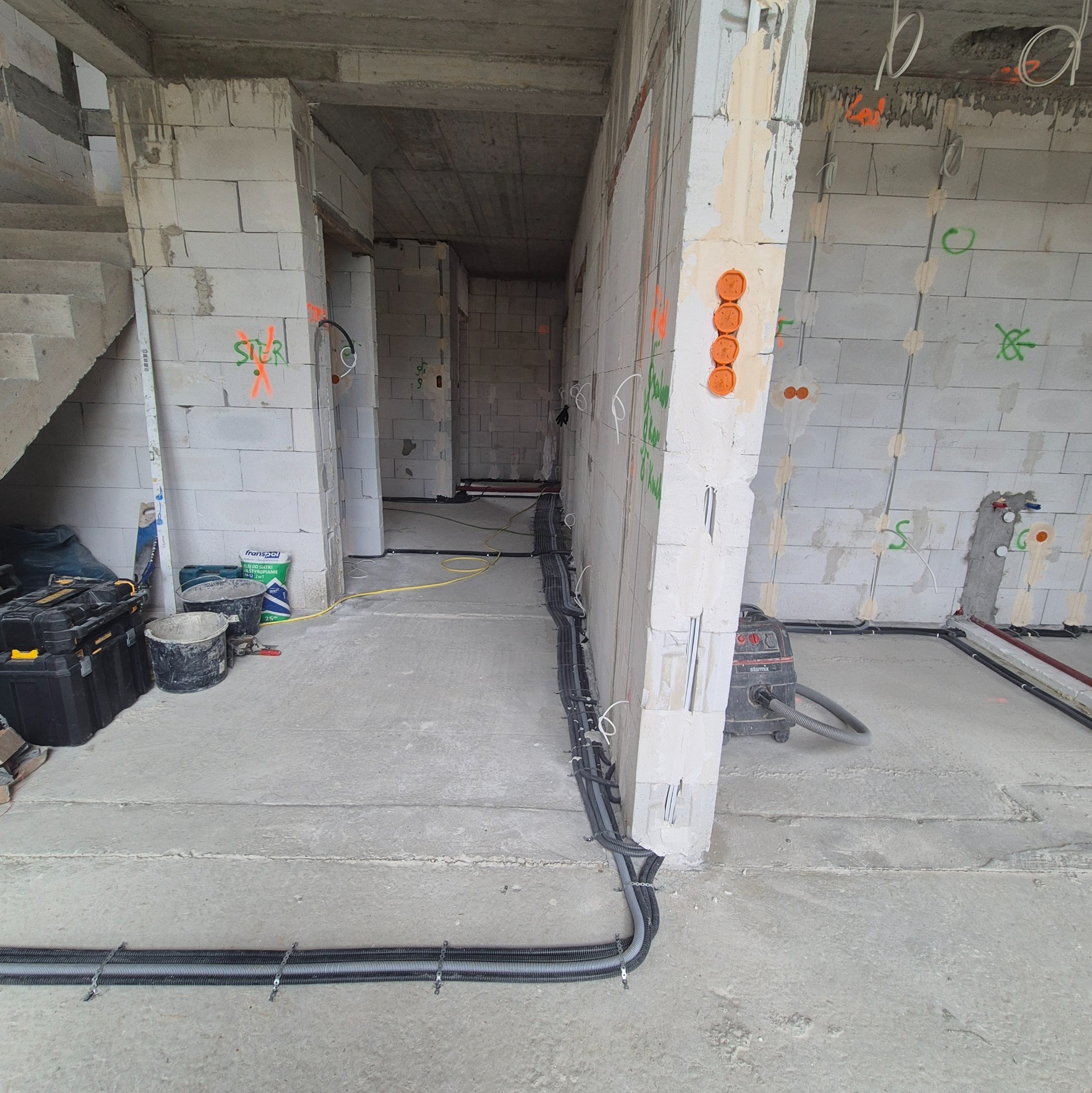 Surowy stan: kable elektryczne w peszlach ułożone na podłodze w budynku w trakcie budowy. Widoczne puszki elektryczne w ścianie i narzędzia.