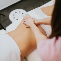Fizjoterapia Dna Miednicy Angelika Gawlik - Masaż pleców wykonywany dłońmi na białym prześcieradle, widok z góry. Terapeuta w różowej bluzce. Białe krzesło w tle. Delikatne oświetlenie.