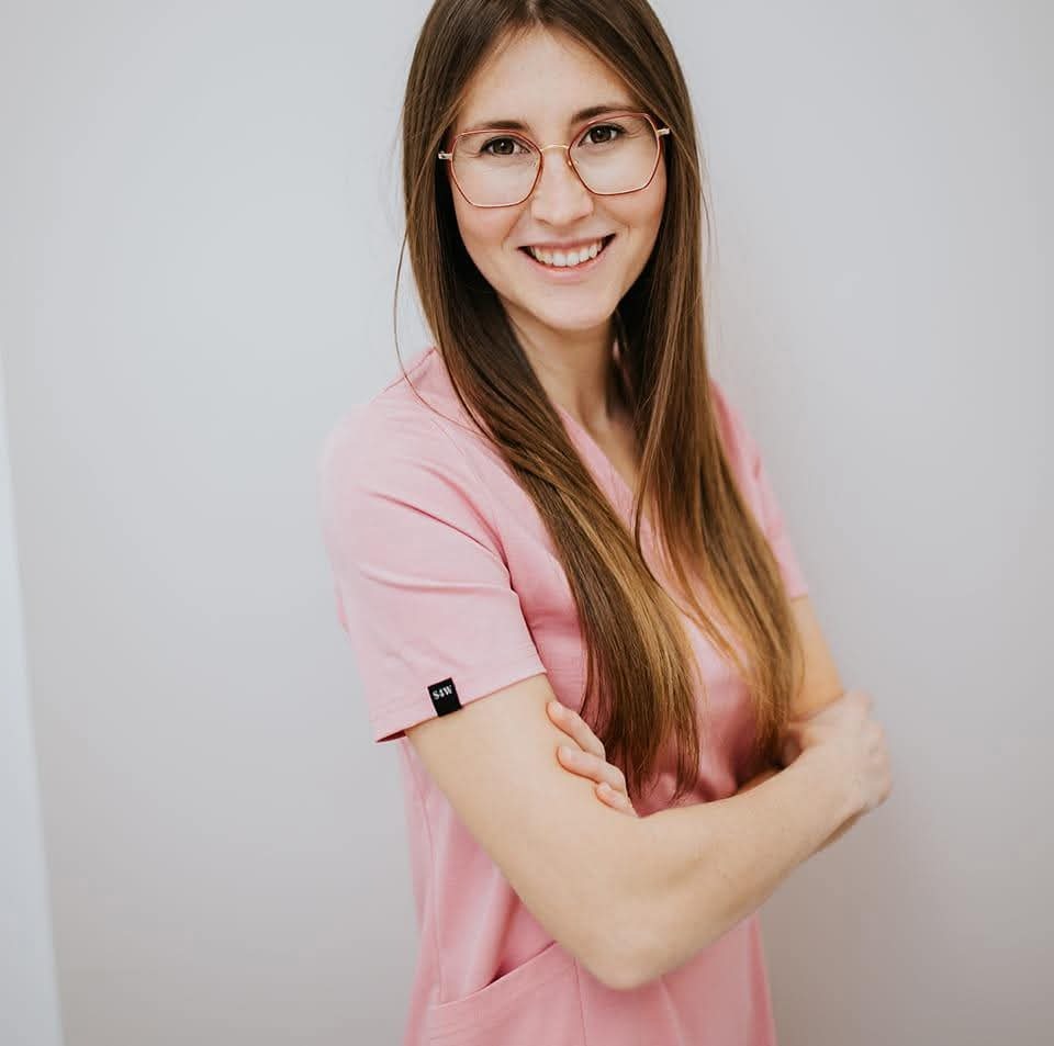 Uśmiechnięta kobieta w różowym uniformie i okularach, z założonymi rękami na tle jasnej ściany. Profesjonalny wizerunek pracownika służby zdrowia.