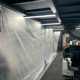 Arkadiusz Burzyński - Biurowe wnętrze zabezpieczone folią malarską przed pracami remontowymi. Widoczne meble biurowe i oświetlenie sufitowe. Przygotowanie do malowania ścian.