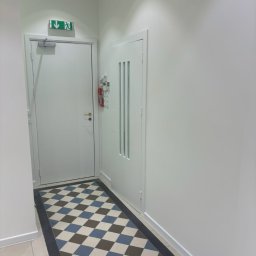 Arkadiusz Burzyński - Białe wnętrze korytarza z wzorzystą podłogą w szachownicę (brąz, błękit, biel), białe ściany i drzwi. Widoczny fragment sufitu z oświetleniem i kamerą.