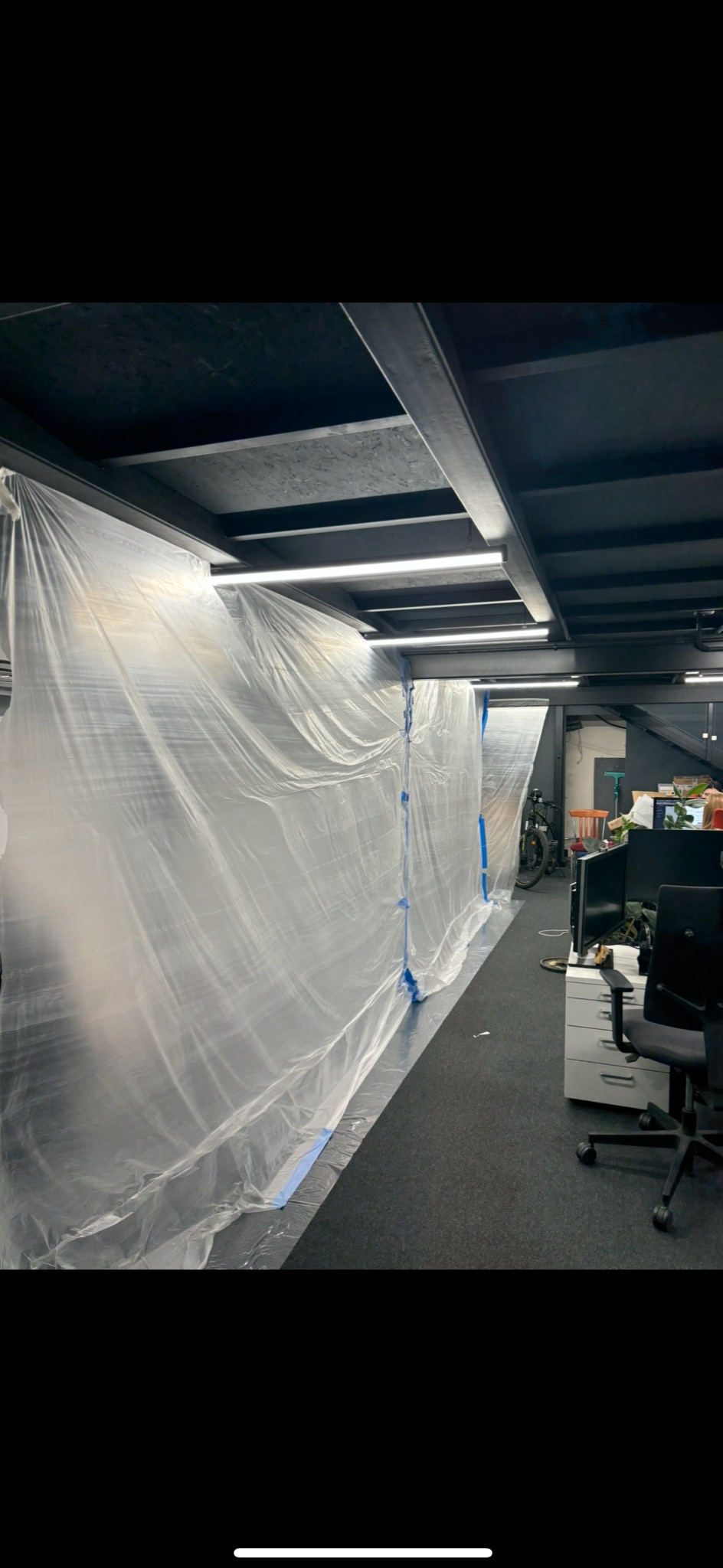 Biurowe wnętrze zabezpieczone folią malarską przed pracami remontowymi. Widoczne meble biurowe i oświetlenie sufitowe. Przygotowanie do malowania ścian.
