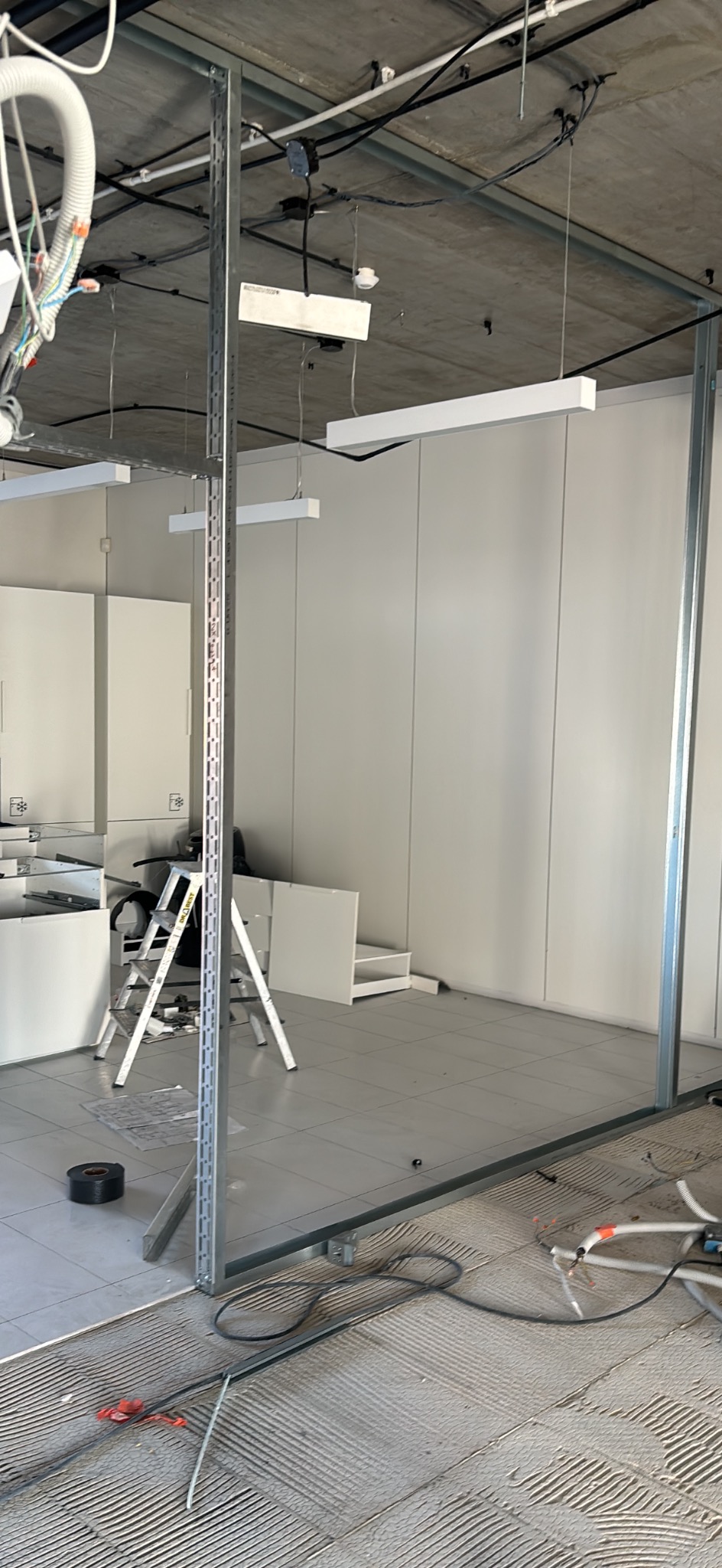 Montaż ścianki działowej z profili metalowych w biurze. Widoczne kable, drabina i elementy wyposażenia. Podłoże przygotowane pod układanie płytek.